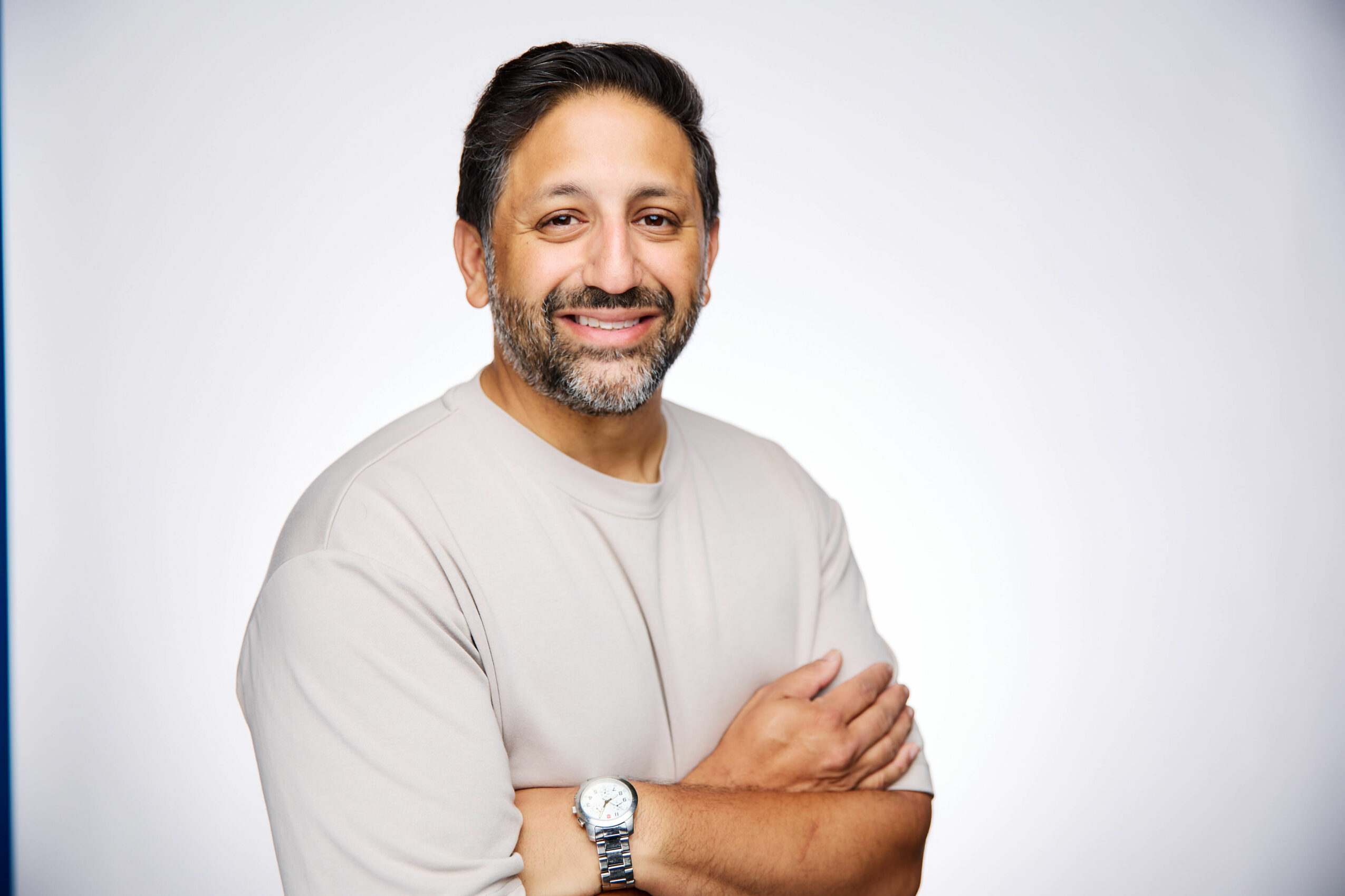 Smiling man with dark hair and beard, wearing a light-colored shirt, crossing arms, against a plain background.