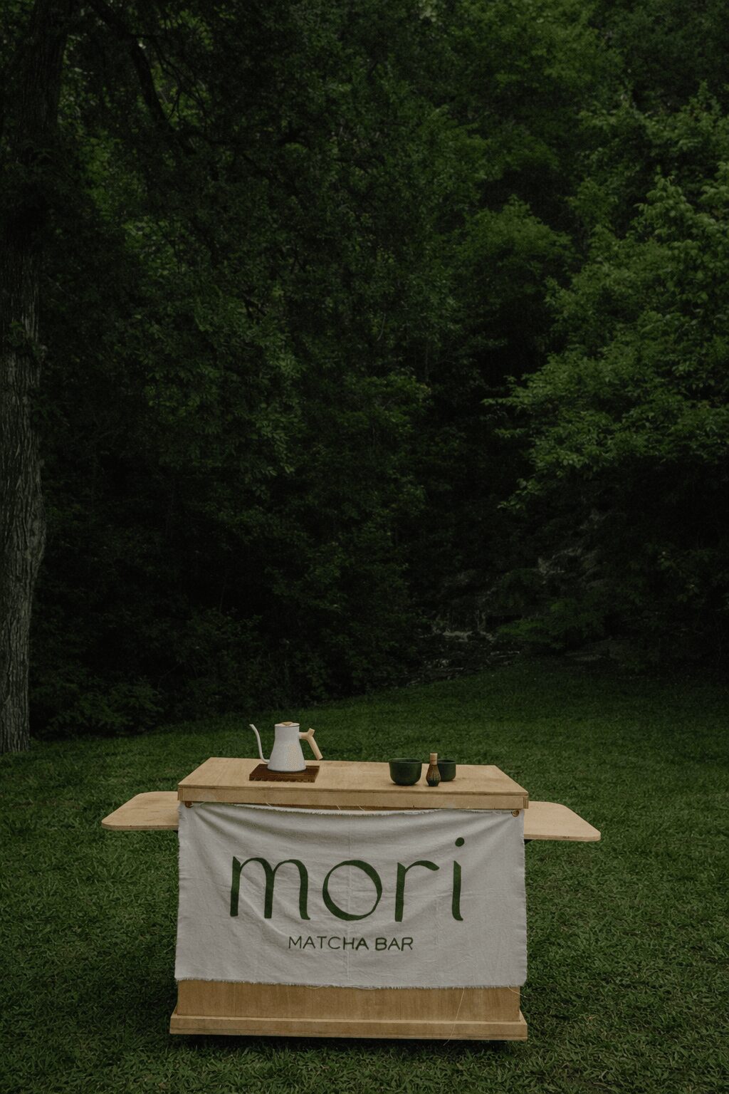 Table with a banner reading 'mori MATCHA BAR' set outdoors in a grassy area with trees.