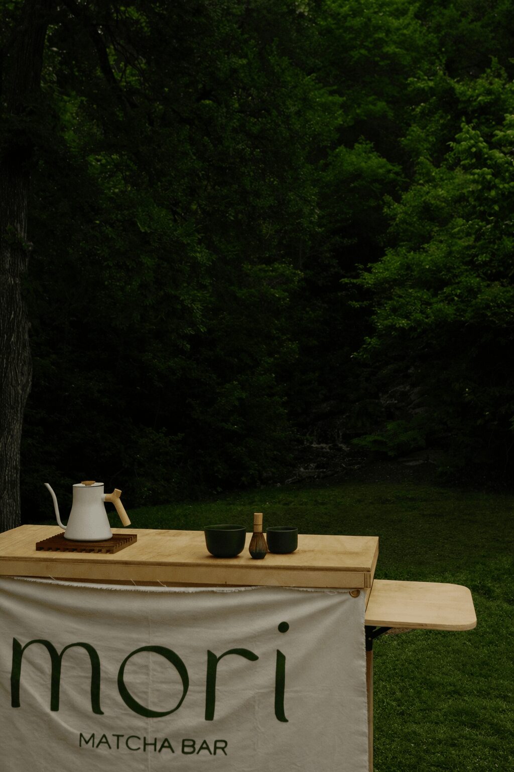 Outdoor table with tea set and banner in a green, wooded area.