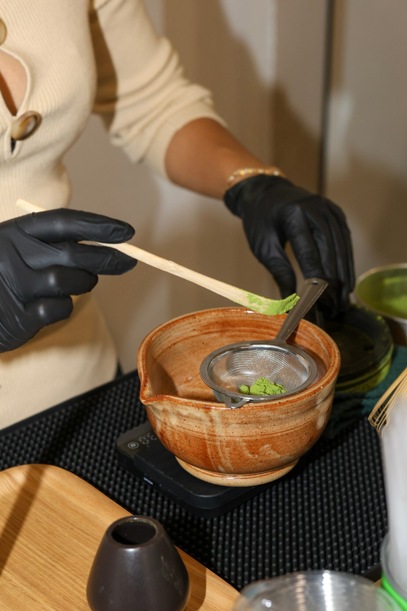 Person wearing black gloves uses a brush to apply green substance into a glass bowl on a wooden table.