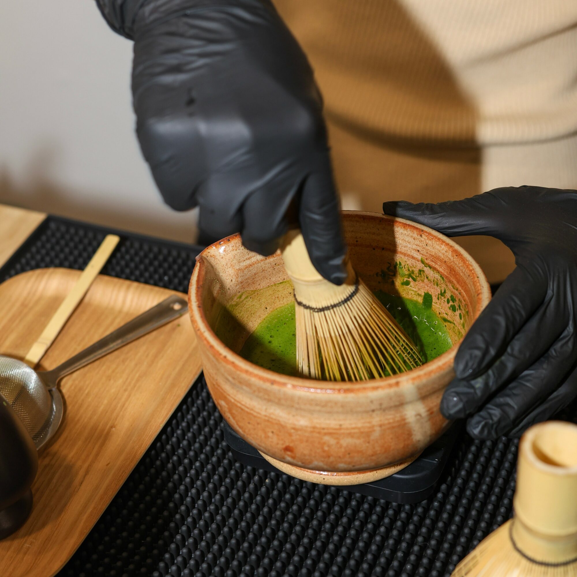 Person whisking green matcha tea in a ceramic bowl with black gloves on a black mat.