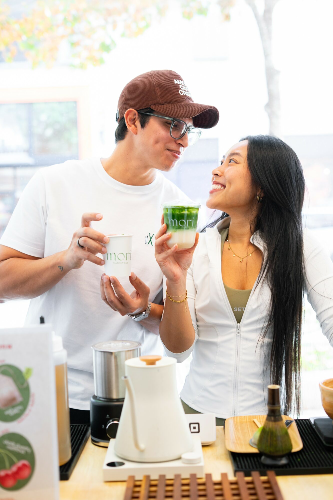 Two people smiling and holding a green cup in a bright kitchen with various items on the counter.