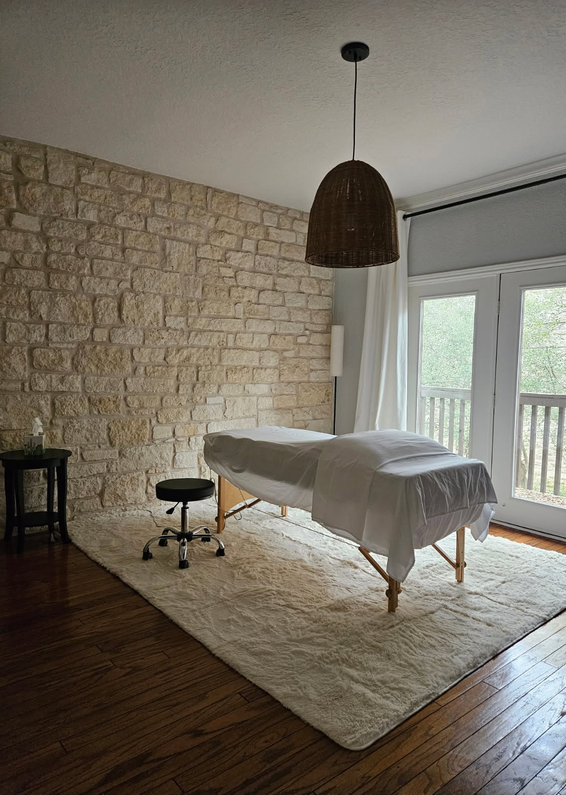Room with stone wall, massage table, stool, and large window with balcony door, natural light, and wooden floor.