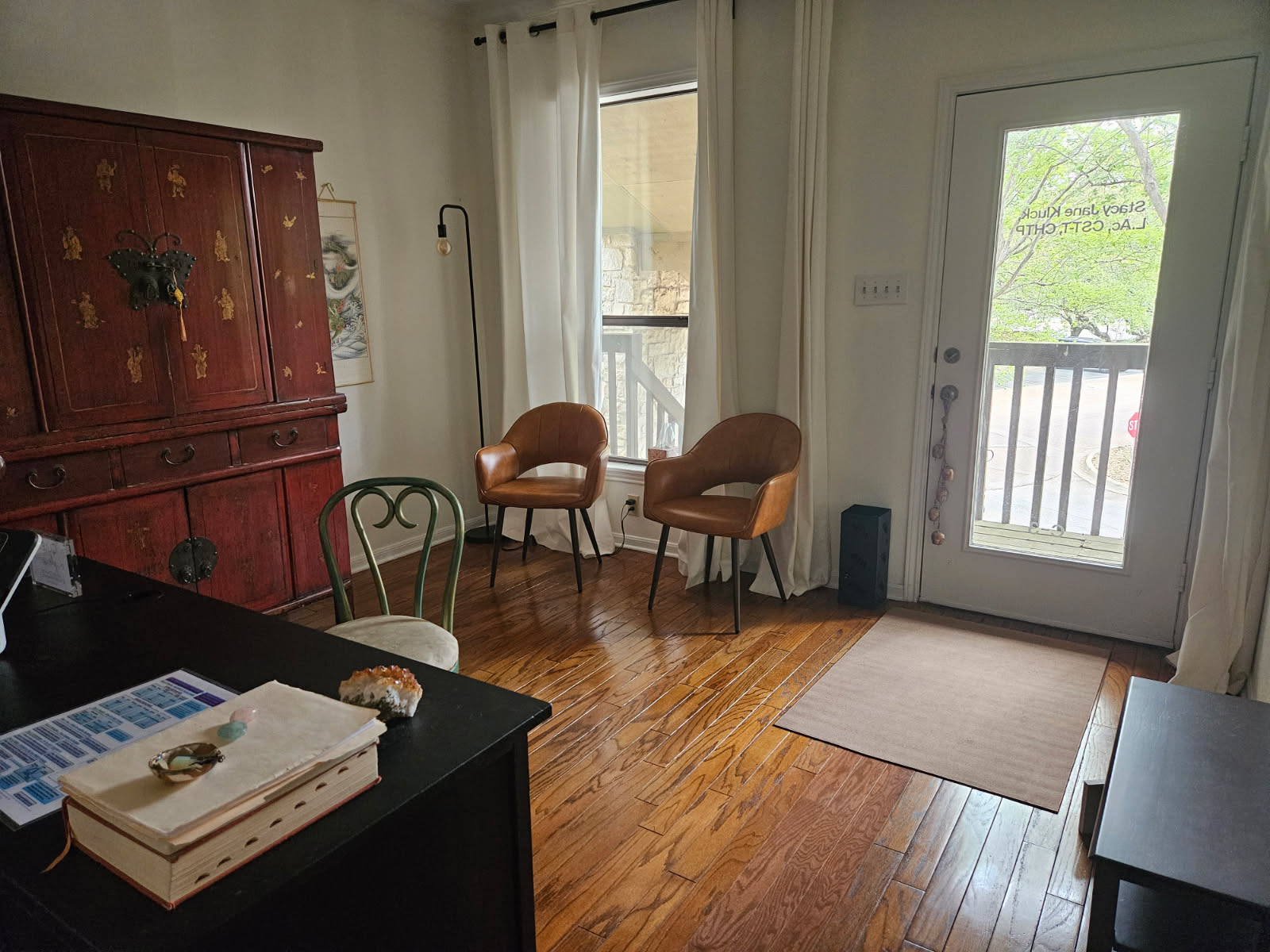 Living room with two chairs near windows, door with balcony, wooden floor, and a cabinet in the corner.