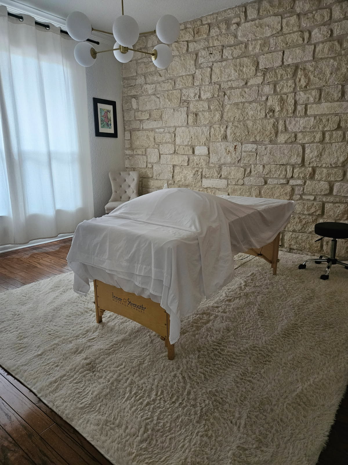 Upright view of a room with a bed covered in white sheets, a stone wall, a window with curtains, and a small black stool.