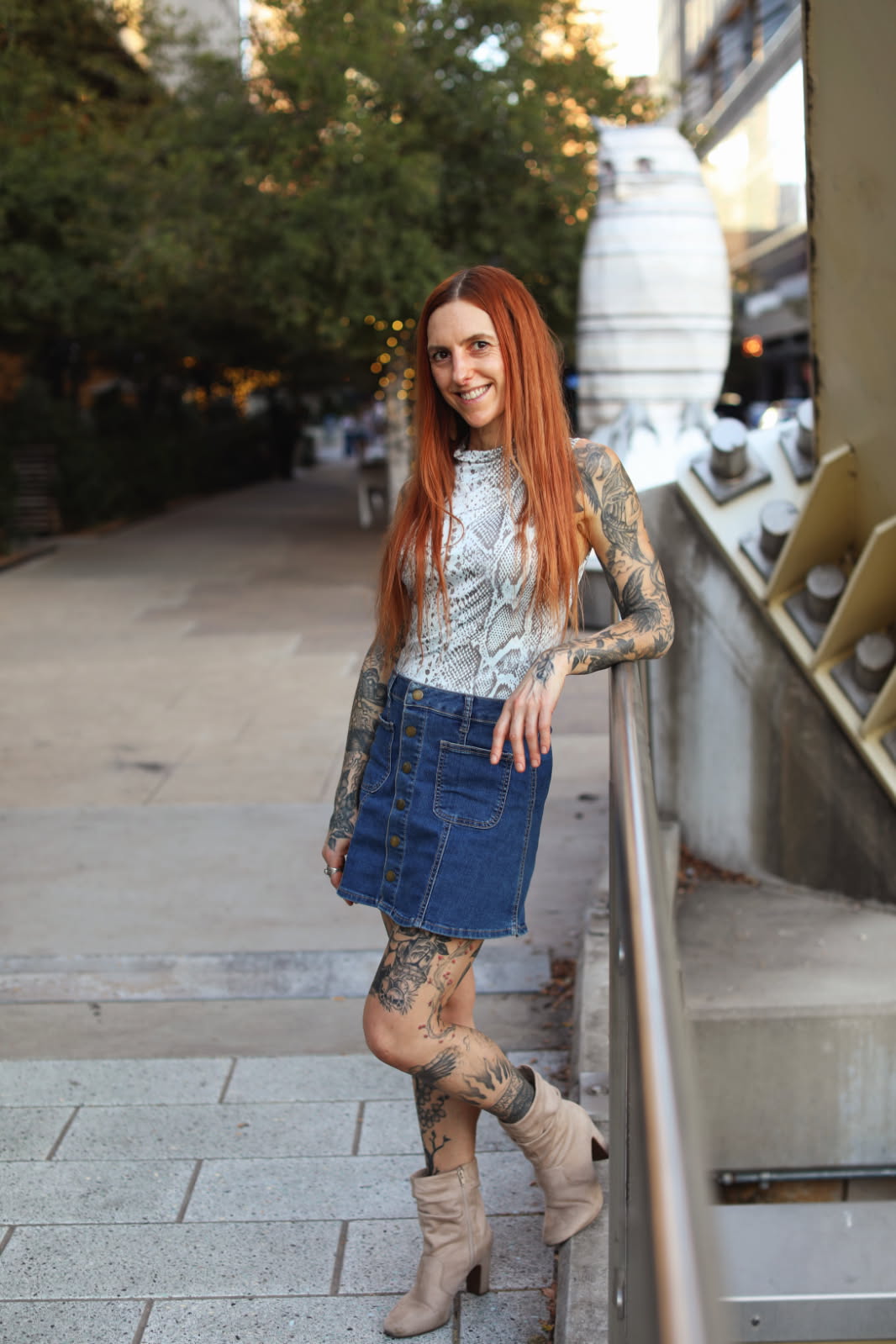 Young woman with long red hair smiling outdoors, leaning on a railing, wearing a lace top, denim skirt, and high-heeled boots.