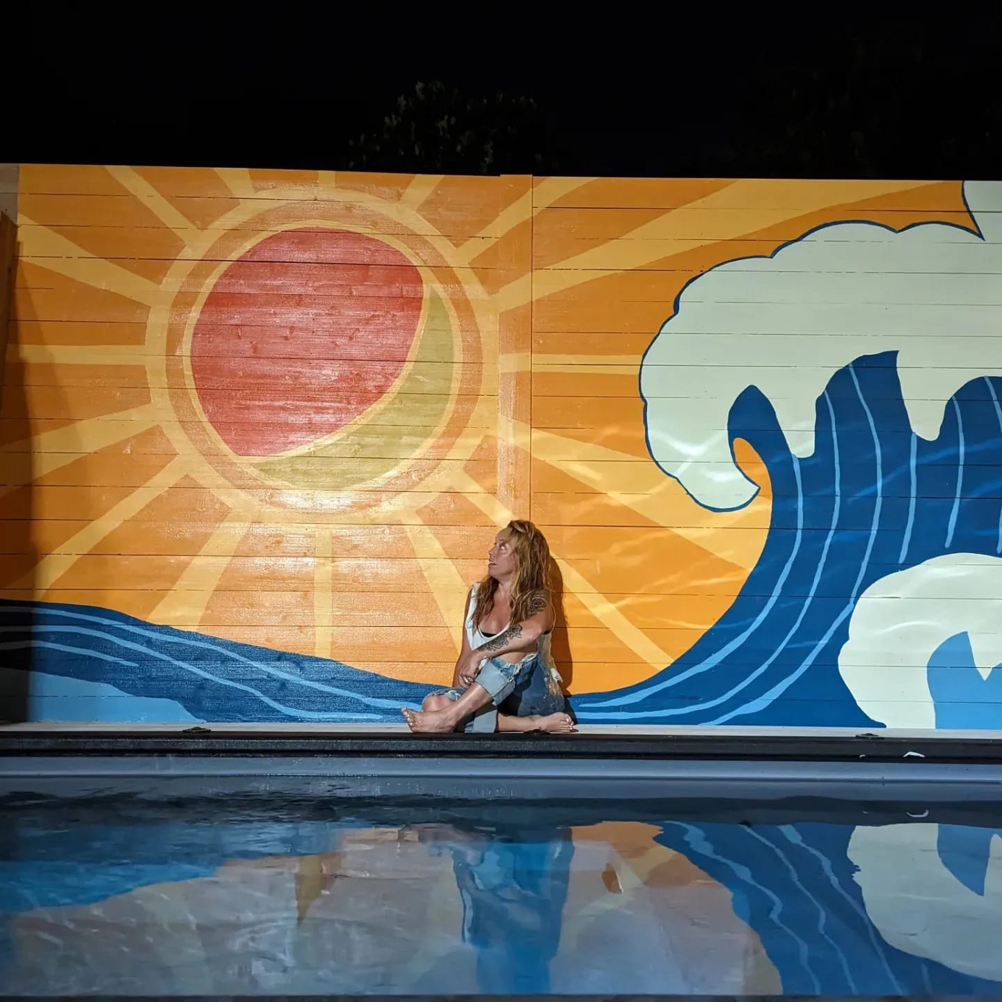Person sitting on pool edge with a colorful mural of a sun and waves behind them.