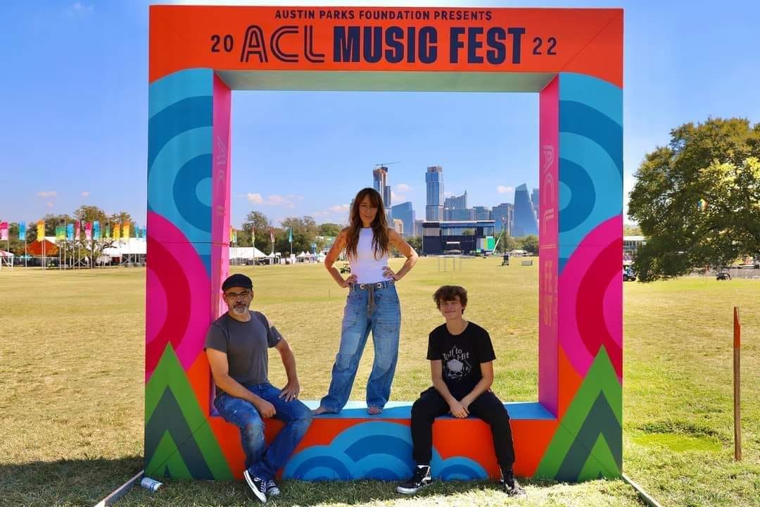 Three people posing in front of a colorful frame at ACL Music Fest 2022, with city skyline and trees in background.