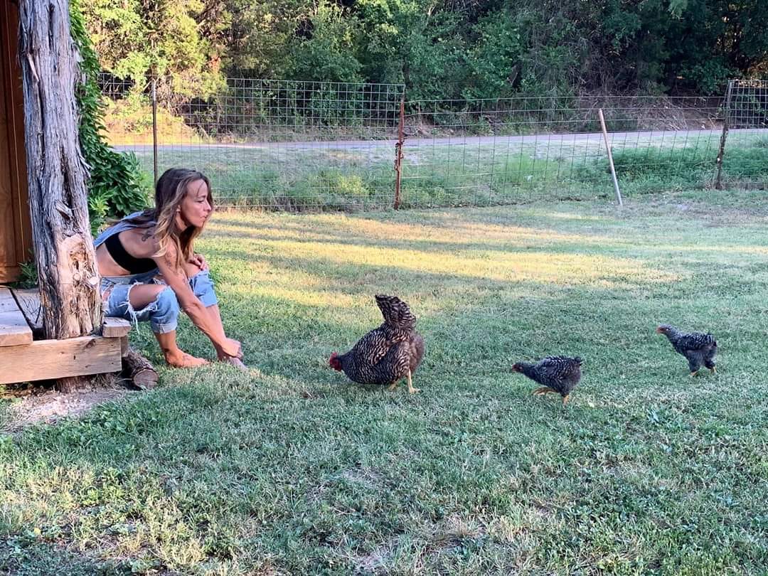 Girl crouching near a tree, playing with three small black puppies on a grassy yard, with a fence and trees in background.