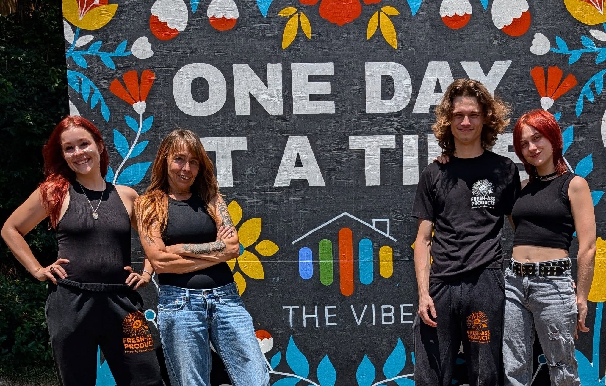 Four people standing in front of a colorful banner with text and floral designs, smiling outdoors.