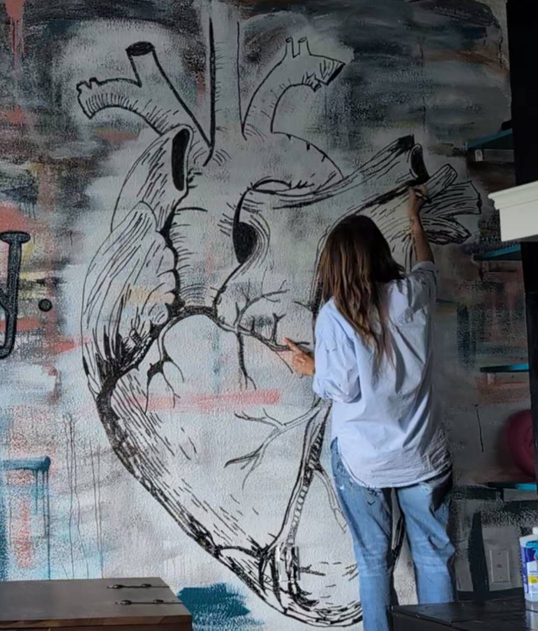 Person with long hair drawing a large anatomical heart on a wall with spray paint, graffiti background, wearing a white shirt and jeans.