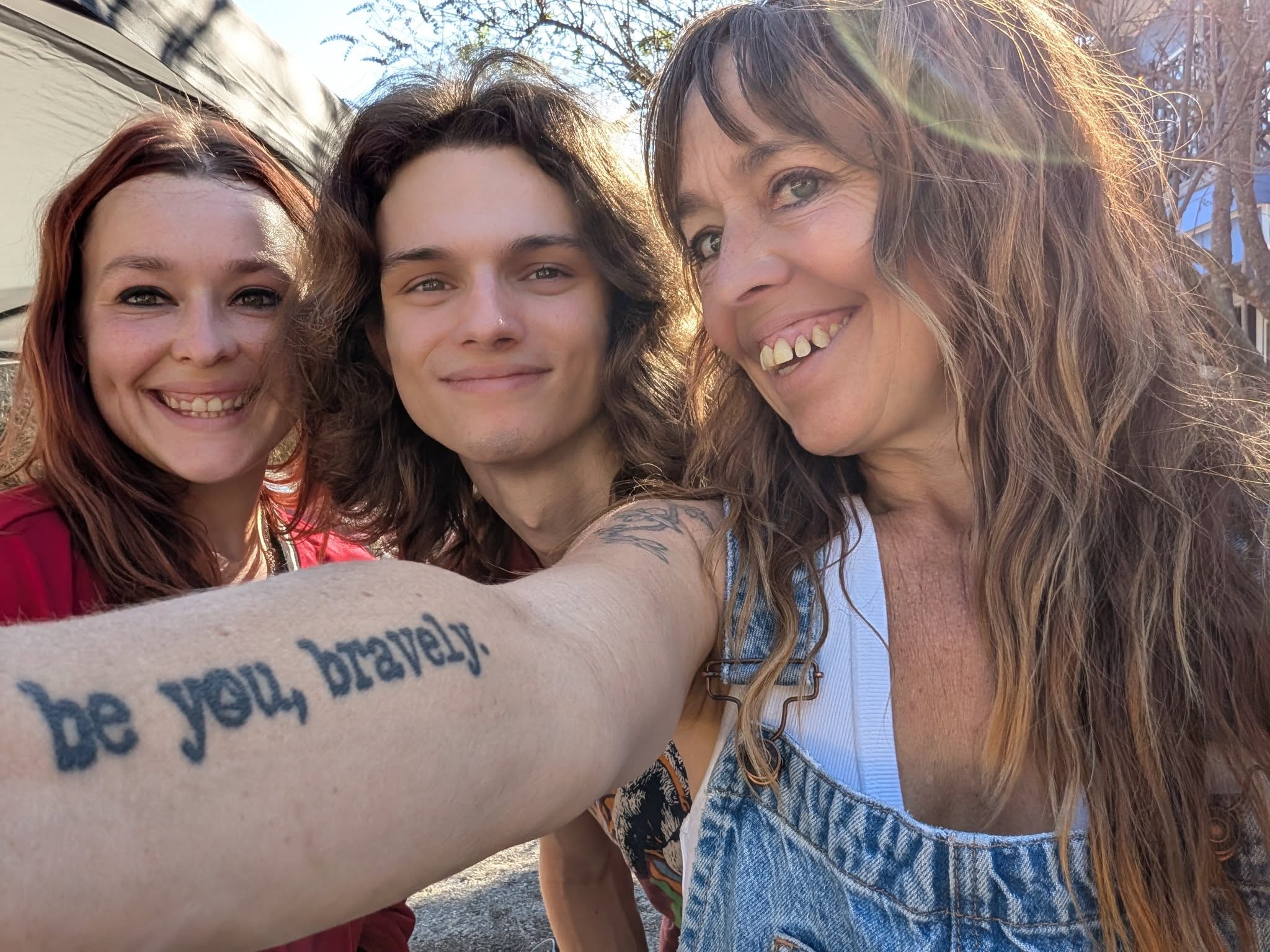 Three smiling women taking a selfie outdoors, with sunlight and trees in the background.