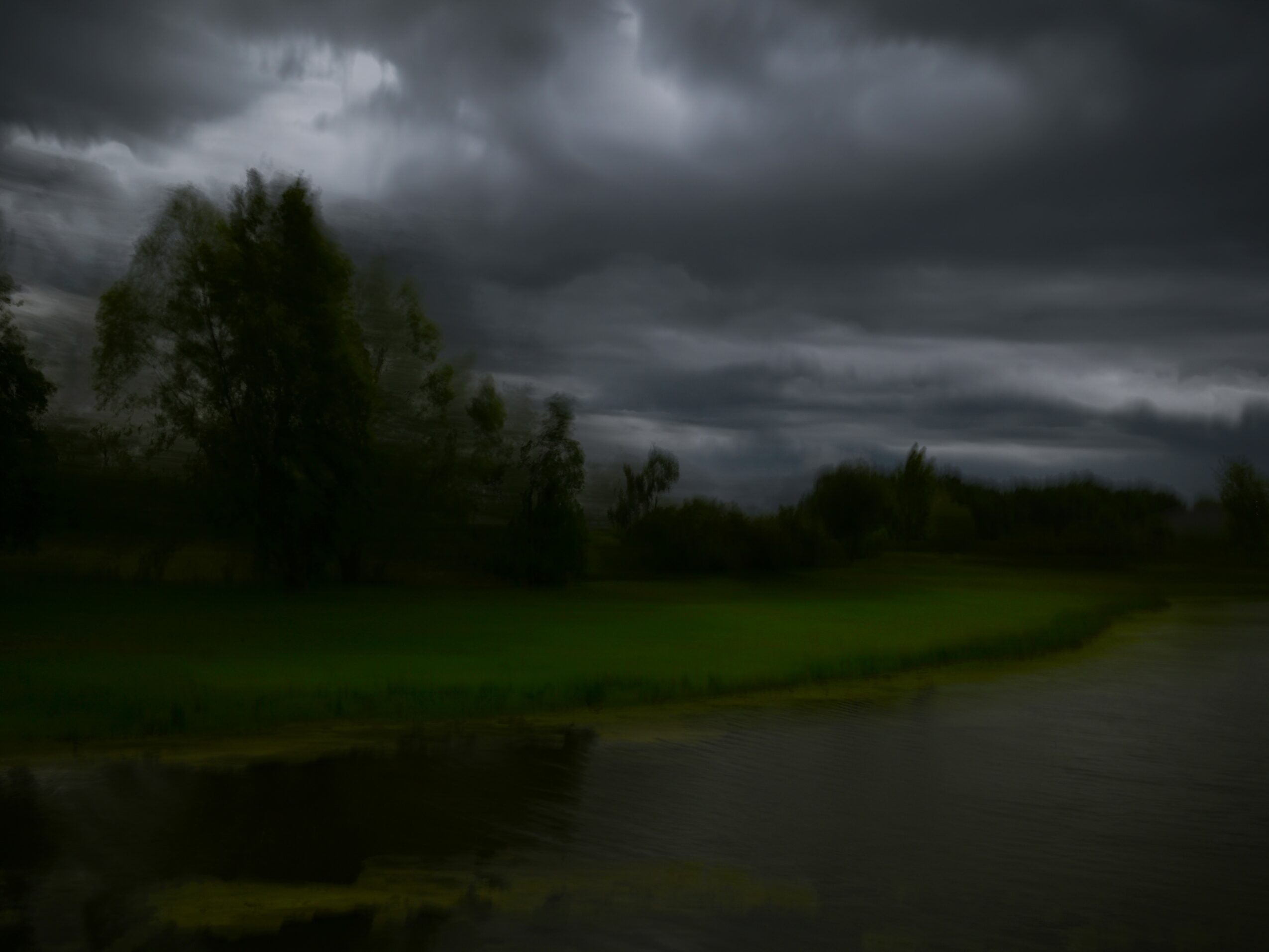 Dark, cloudy sky over a landscape with trees, grass, and a body of water, indicating an approaching storm.