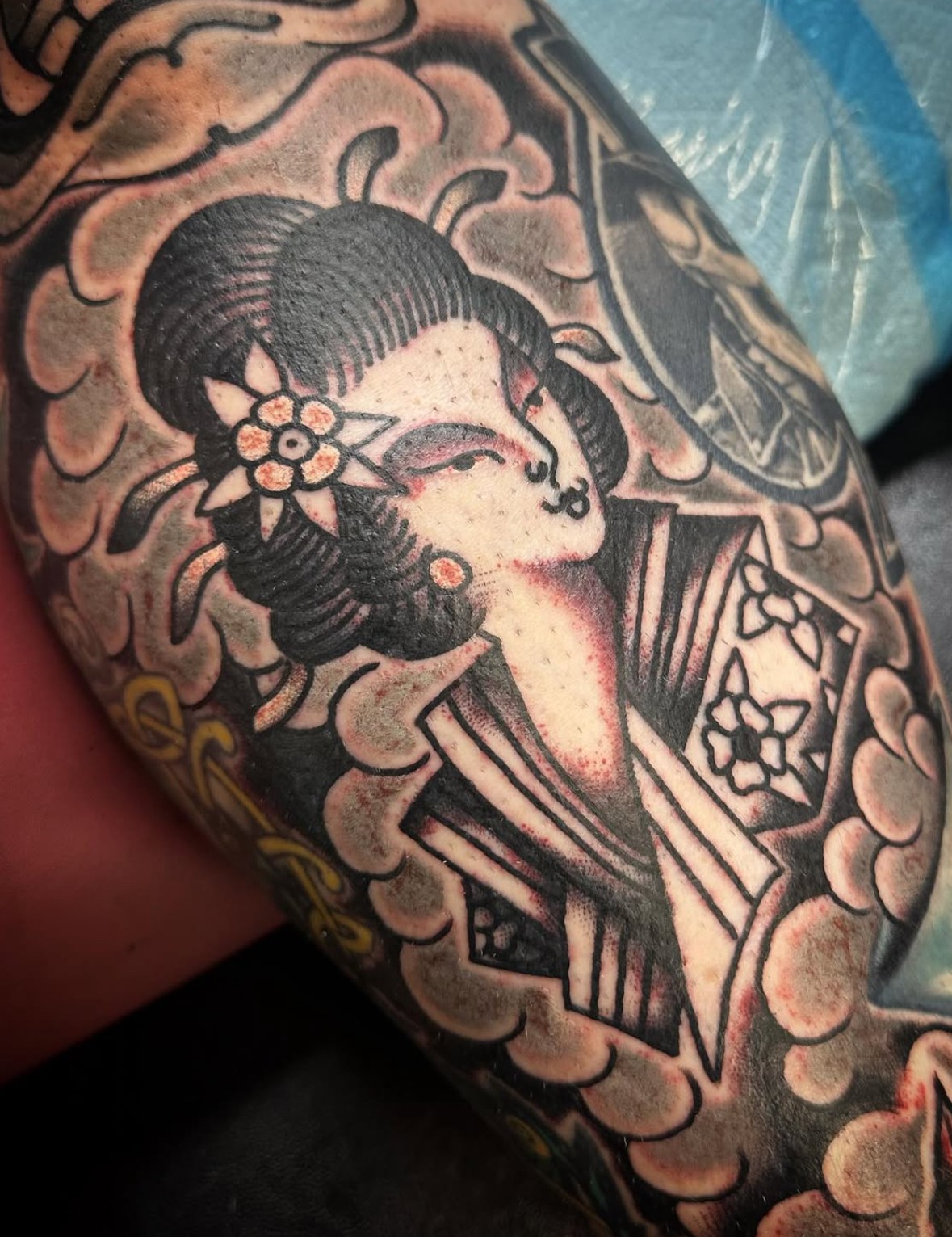Tattoo of a woman with traditional Japanese hairstyle and flowers, surrounded by clouds, in black and gray ink.