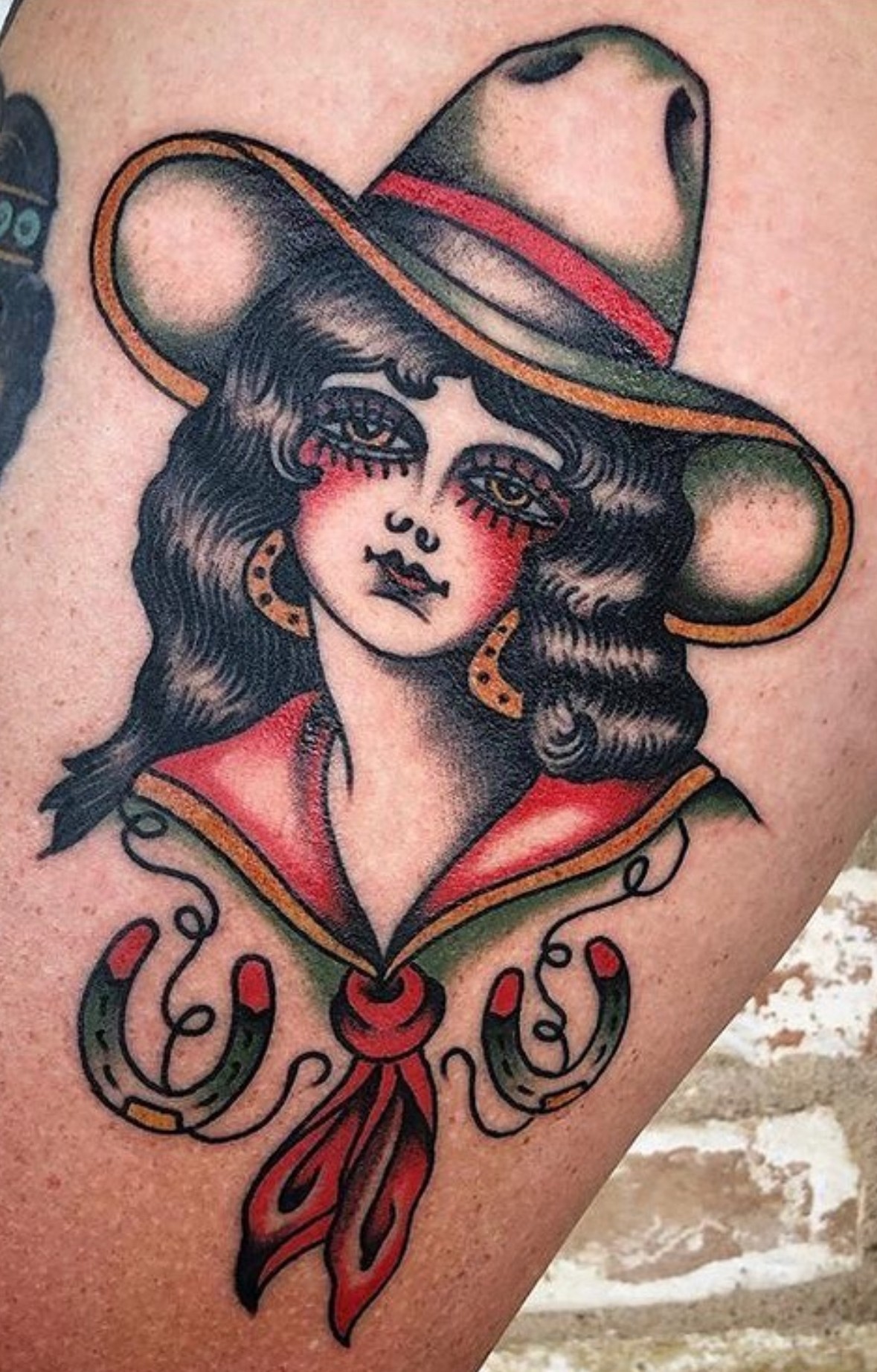 Tattoo of a woman with long dark hair, wearing a wide-brimmed hat, earrings, and a red scarf, with decorative elements below.