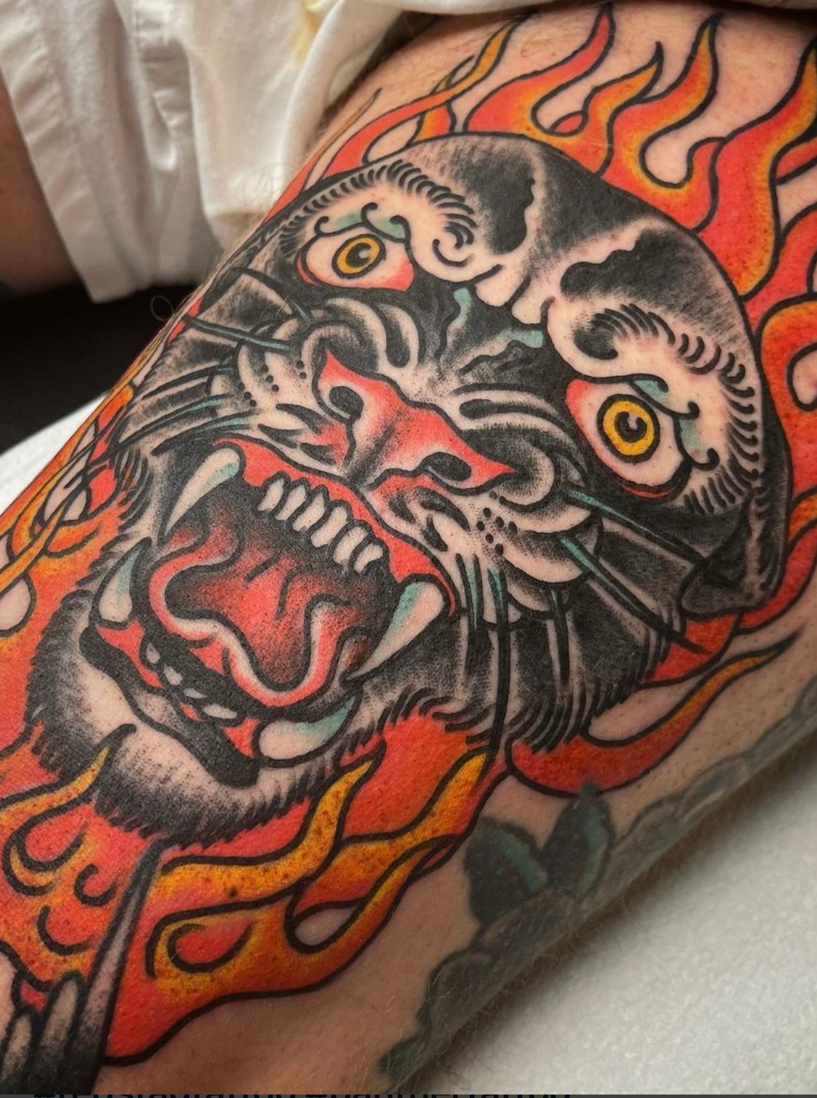 Tattoo of a roaring tiger's face with flames around it, detailed with black, gray, and red colors.