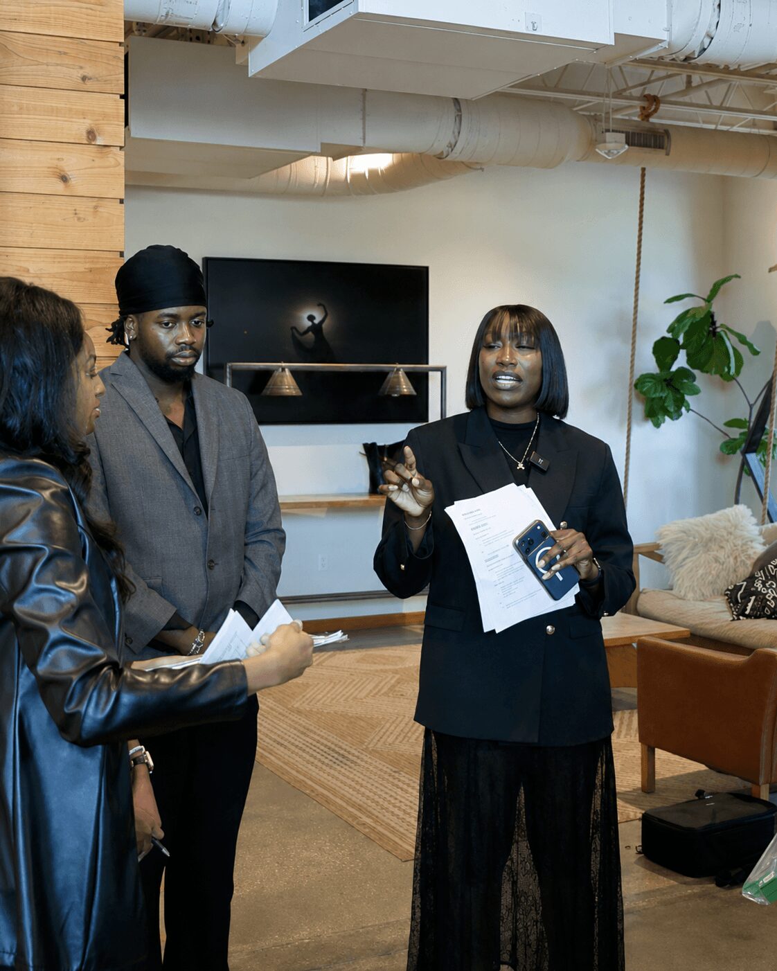 Three people in conversation indoors, one woman speaking and holding papers, two others listening, casual setting with furniture and plants.