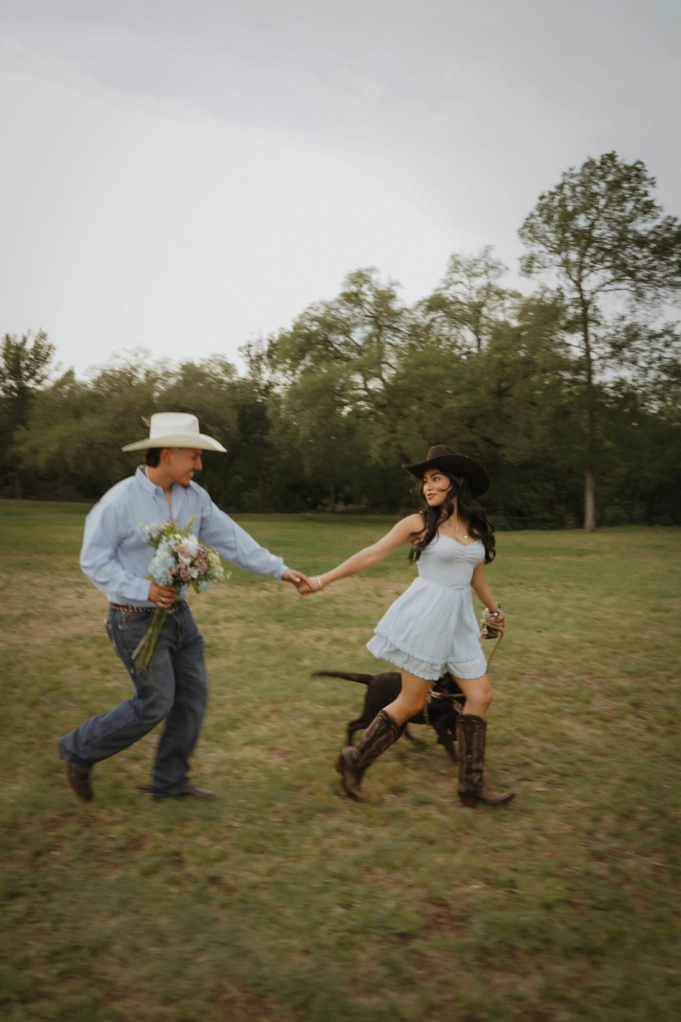 A man and woman holding hands in a grassy field with trees, the man wearing a cowboy hat and jeans, the woman in a white dress and boots.