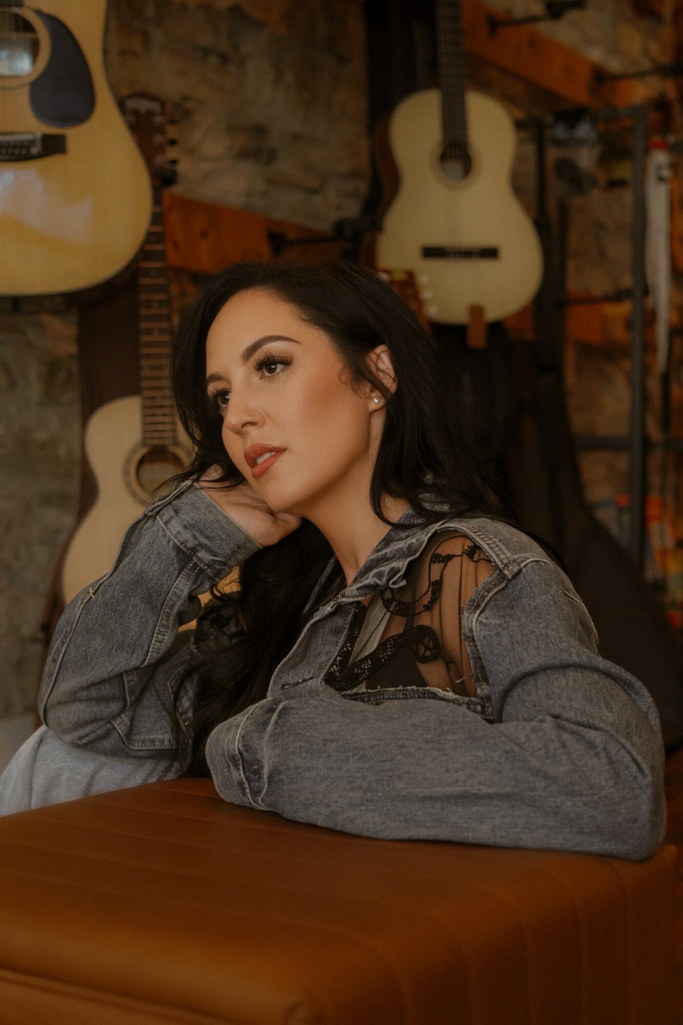 Young woman with dark hair and makeup, wearing a denim jacket, sitting at a table in a guitar shop or studio, with guitars hanging on the wall behind her.