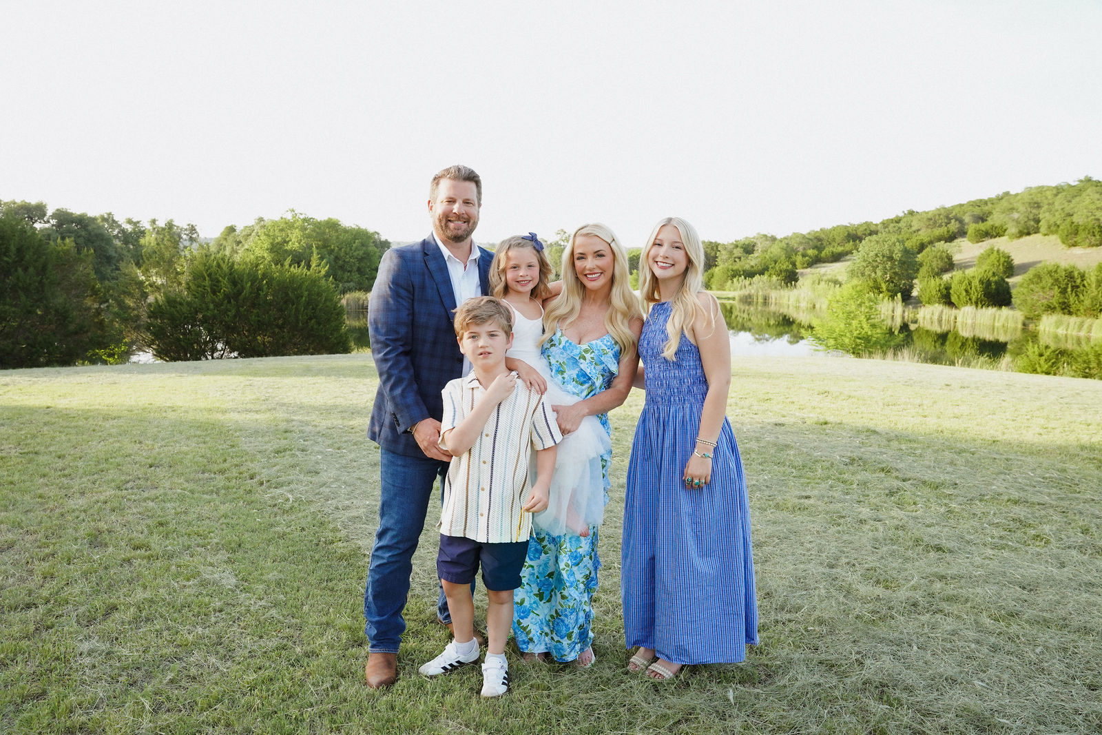 Group of five people standing outdoors on a grassy field with trees and hills in background, smiling, dressed in summer clothes.