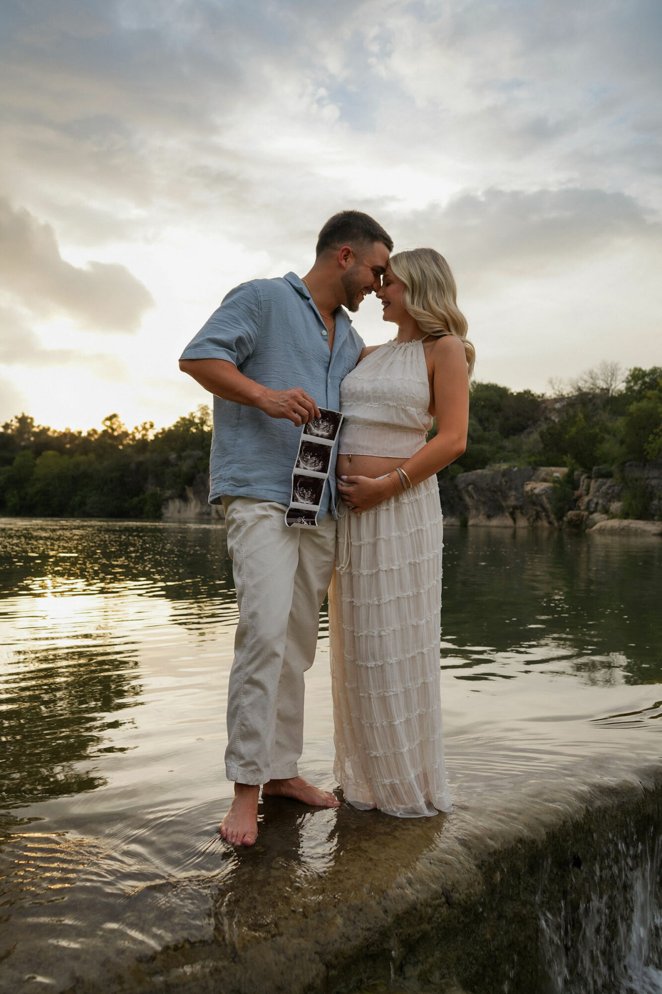 A couple stands in water near a river, embracing and smiling at each other during sunset, with trees in the background.