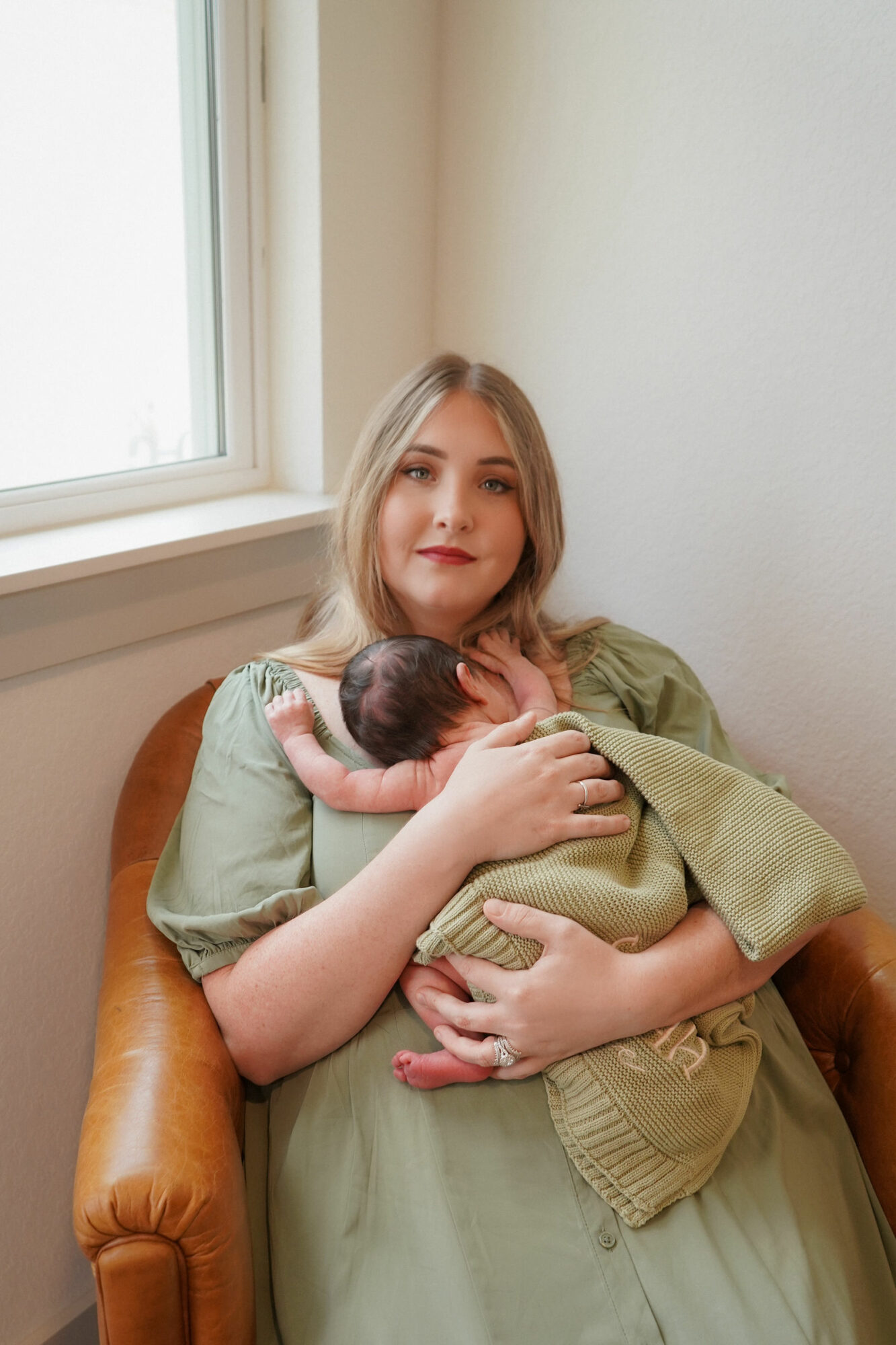 Young woman sitting on a chair holding a newborn baby near a window, with a neutral background.