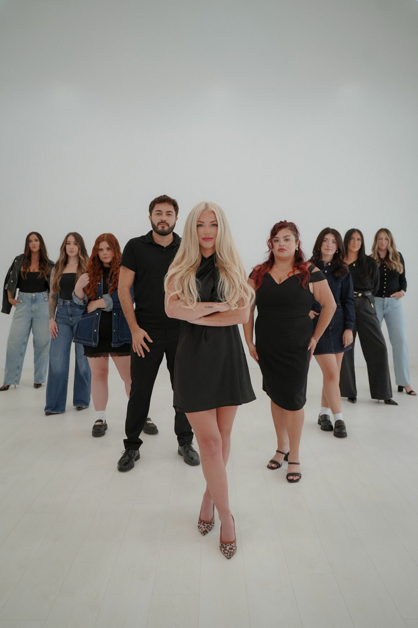 Group of nine women and one man standing in a studio with a plain light background, the woman in front has long blonde hair and crossed arms.