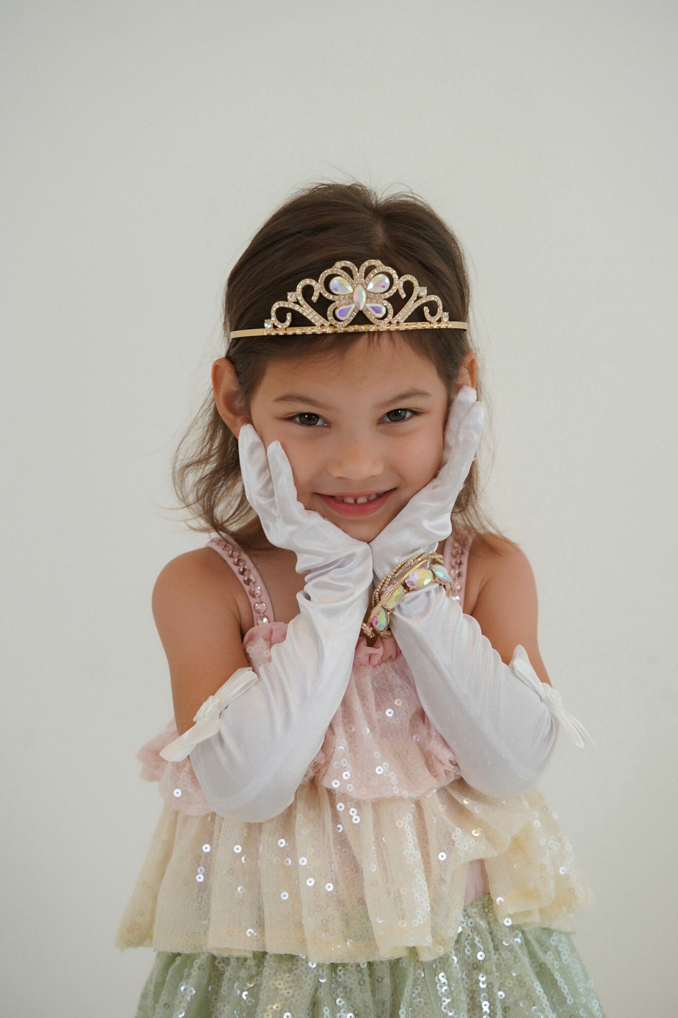 Young girl wearing a tiara and gloves, smiling with hands on cheeks, dressed in a pastel-colored dress, against a plain background.