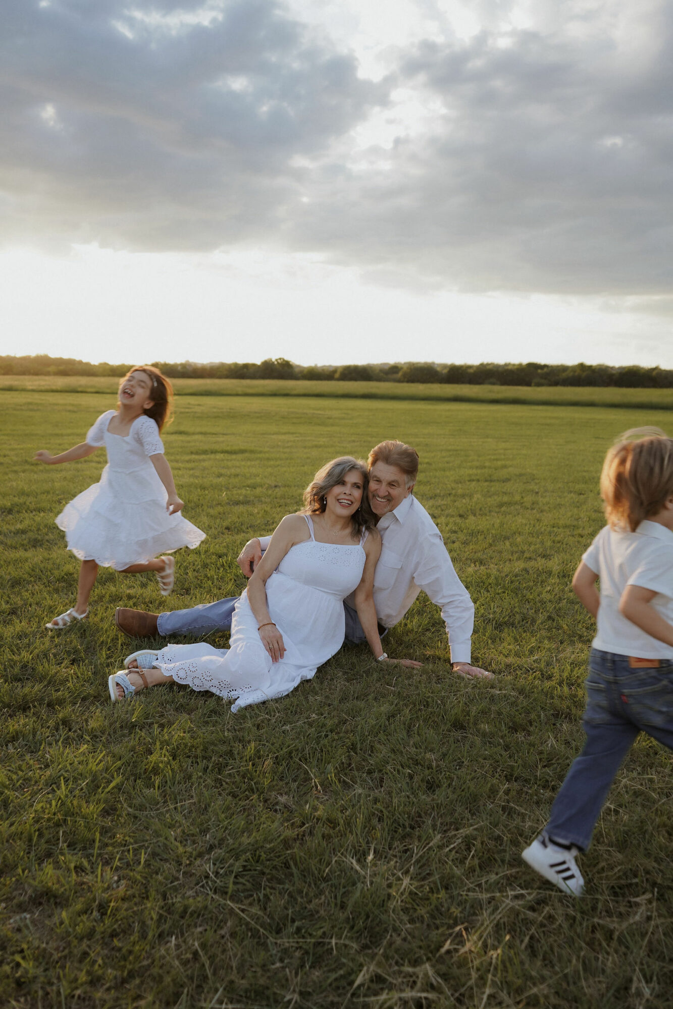 Two women in white dresses sit on grass, smiling, with children playing nearby in an open field under cloudy sky.