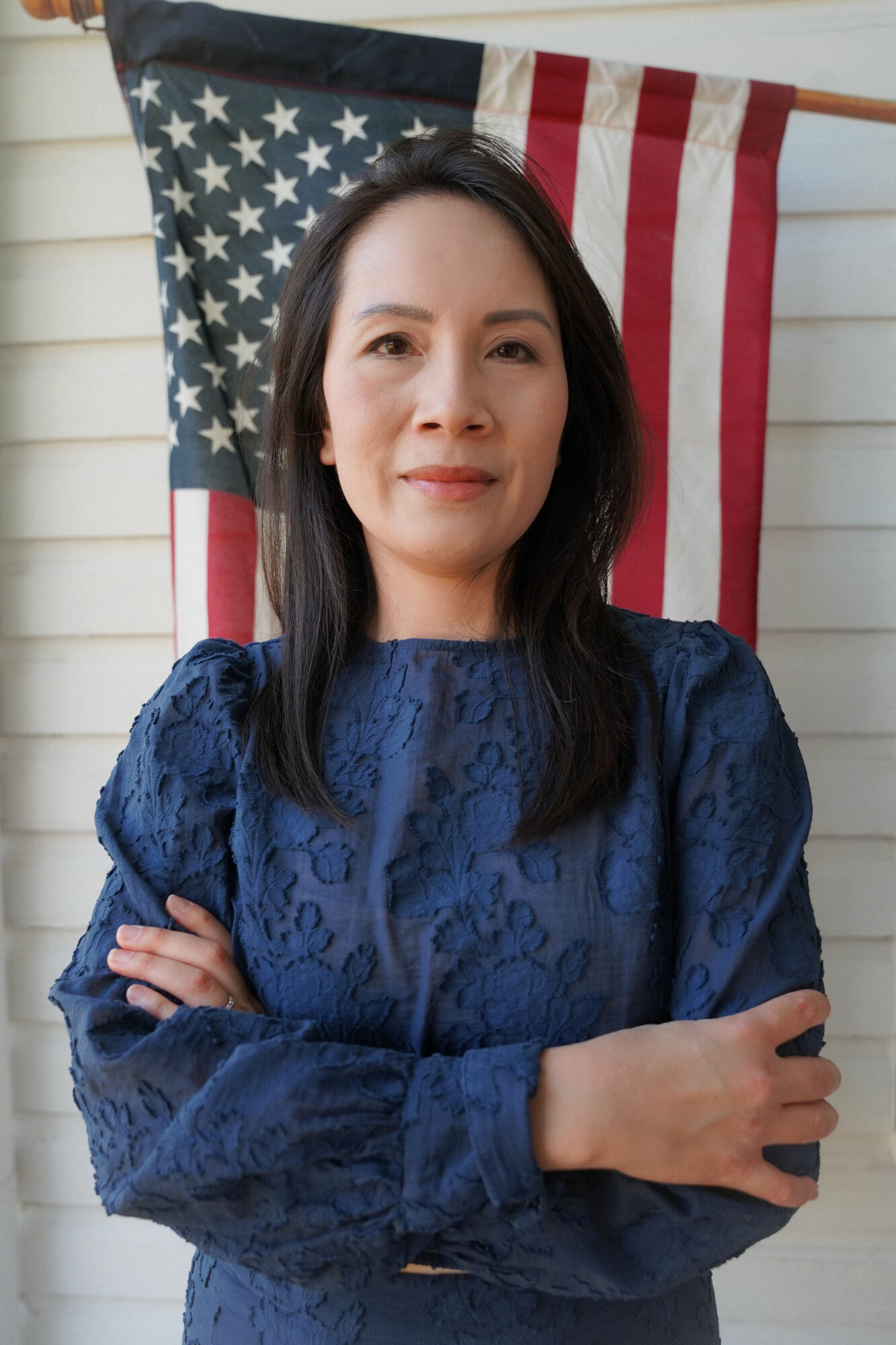 Woman with dark hair in front of an American flag, wearing a blue lace top, arms crossed, smiling.