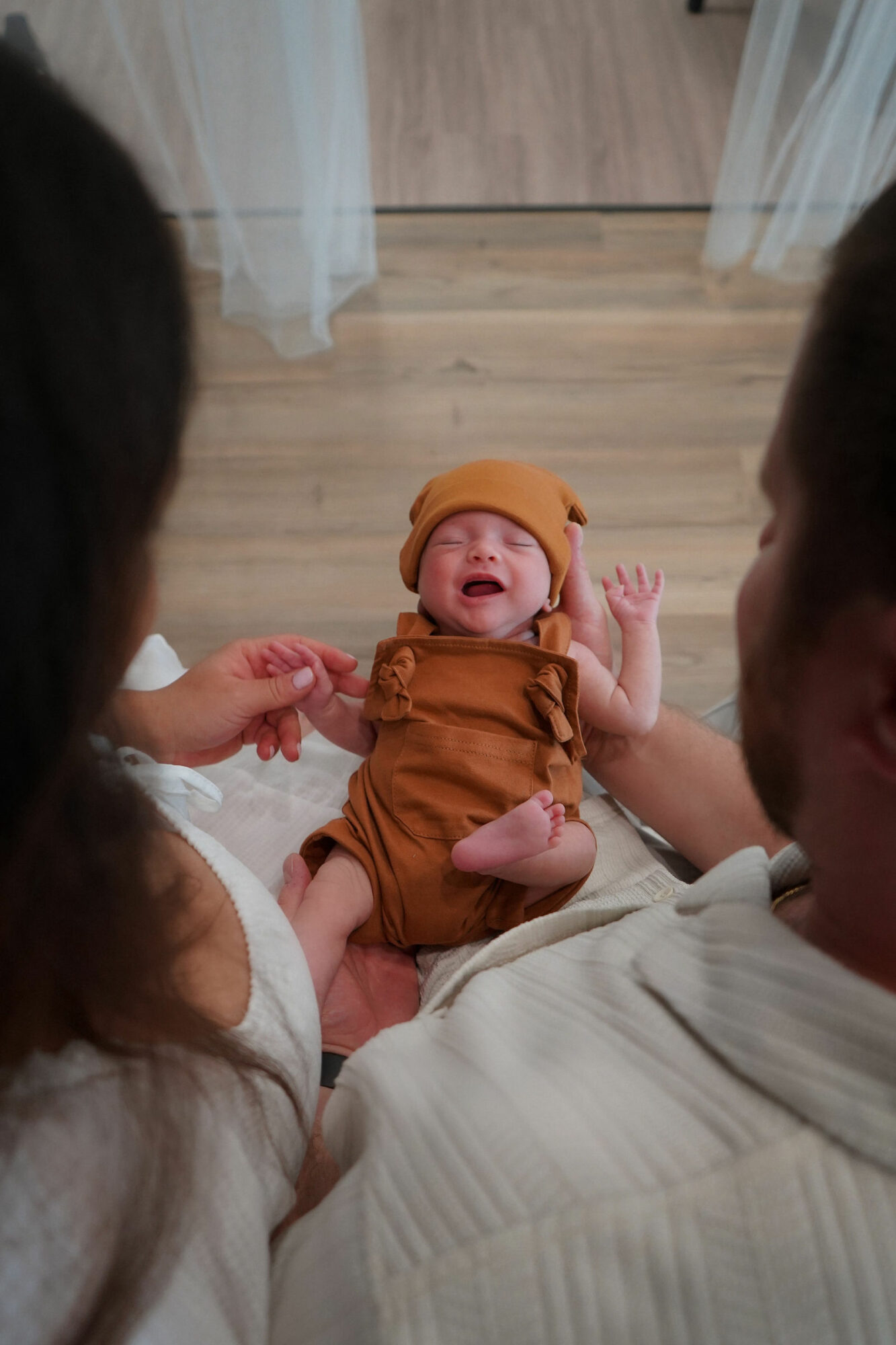 Baby in brown outfit and hat crying, held by two adults, on wooden floor with curtains in background.