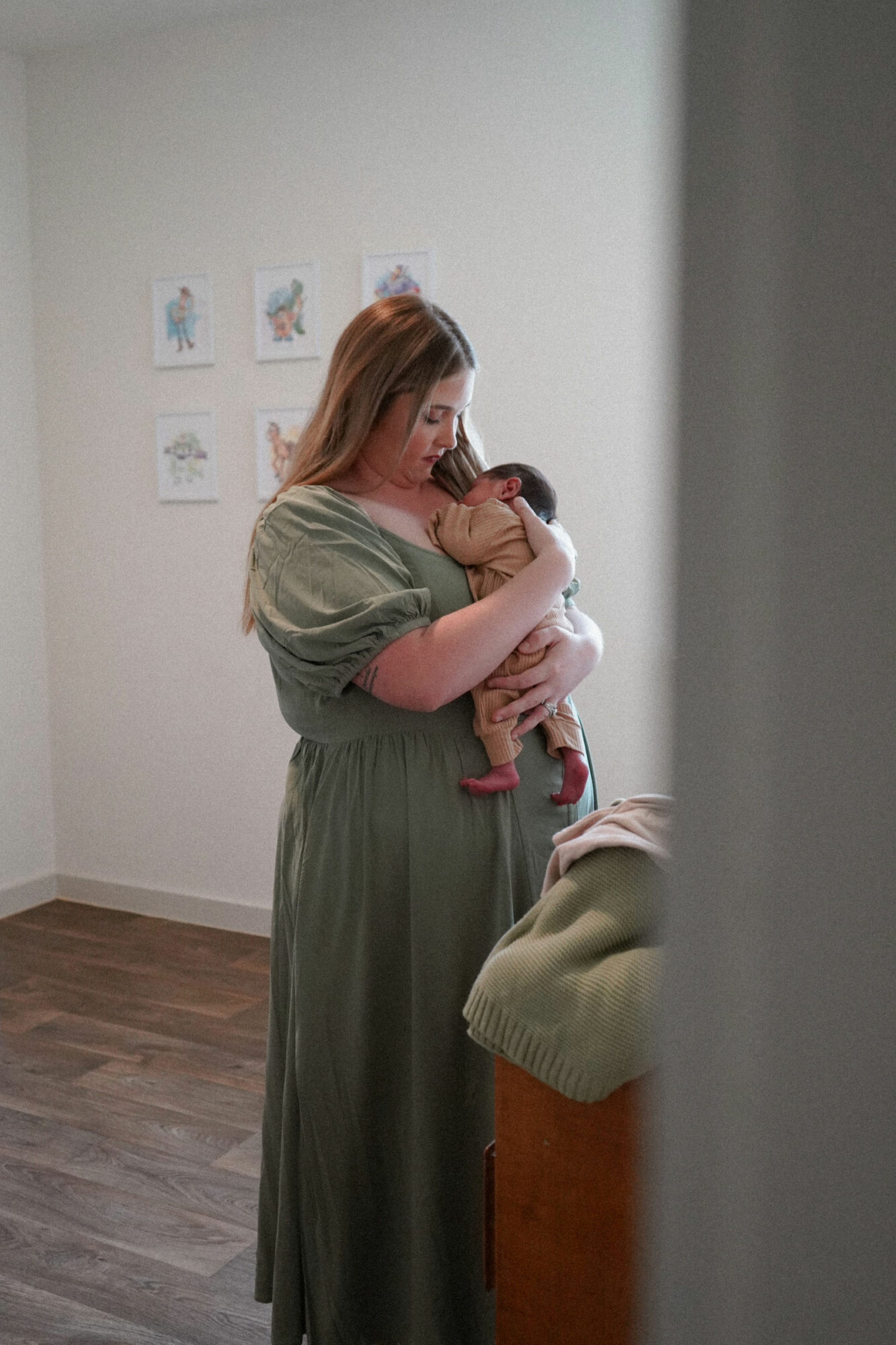 Woman holding a baby in a room with wall art, standing near a piece of furniture, soft lighting.