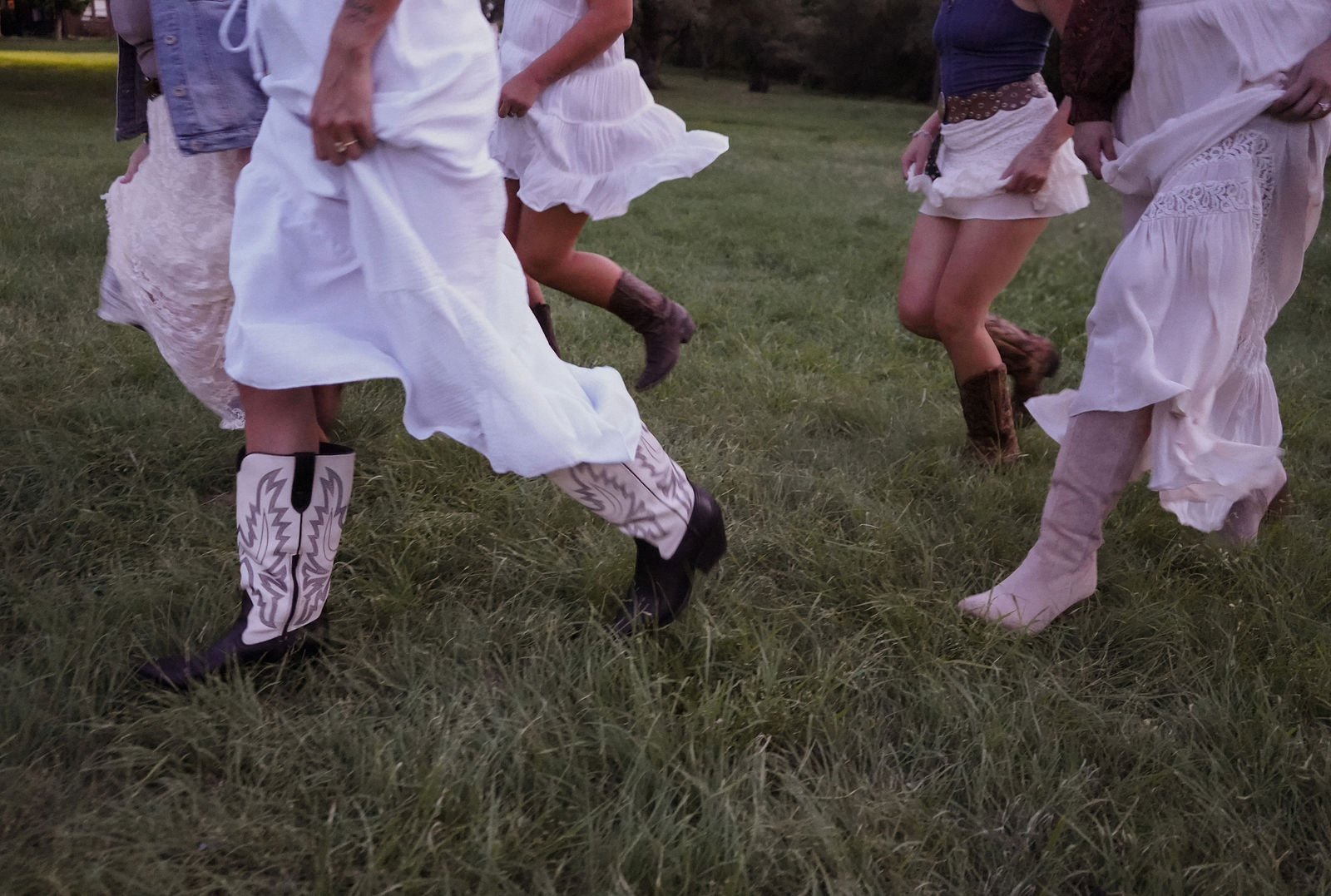 Group of people dancing outdoors, focusing on their legs and feet, wearing white and patterned clothing, on grass.