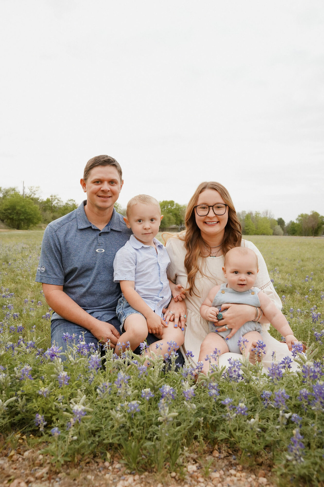 Family of four sitting in a field of purple flowers outdoors, smiling, with trees and sky in background.
