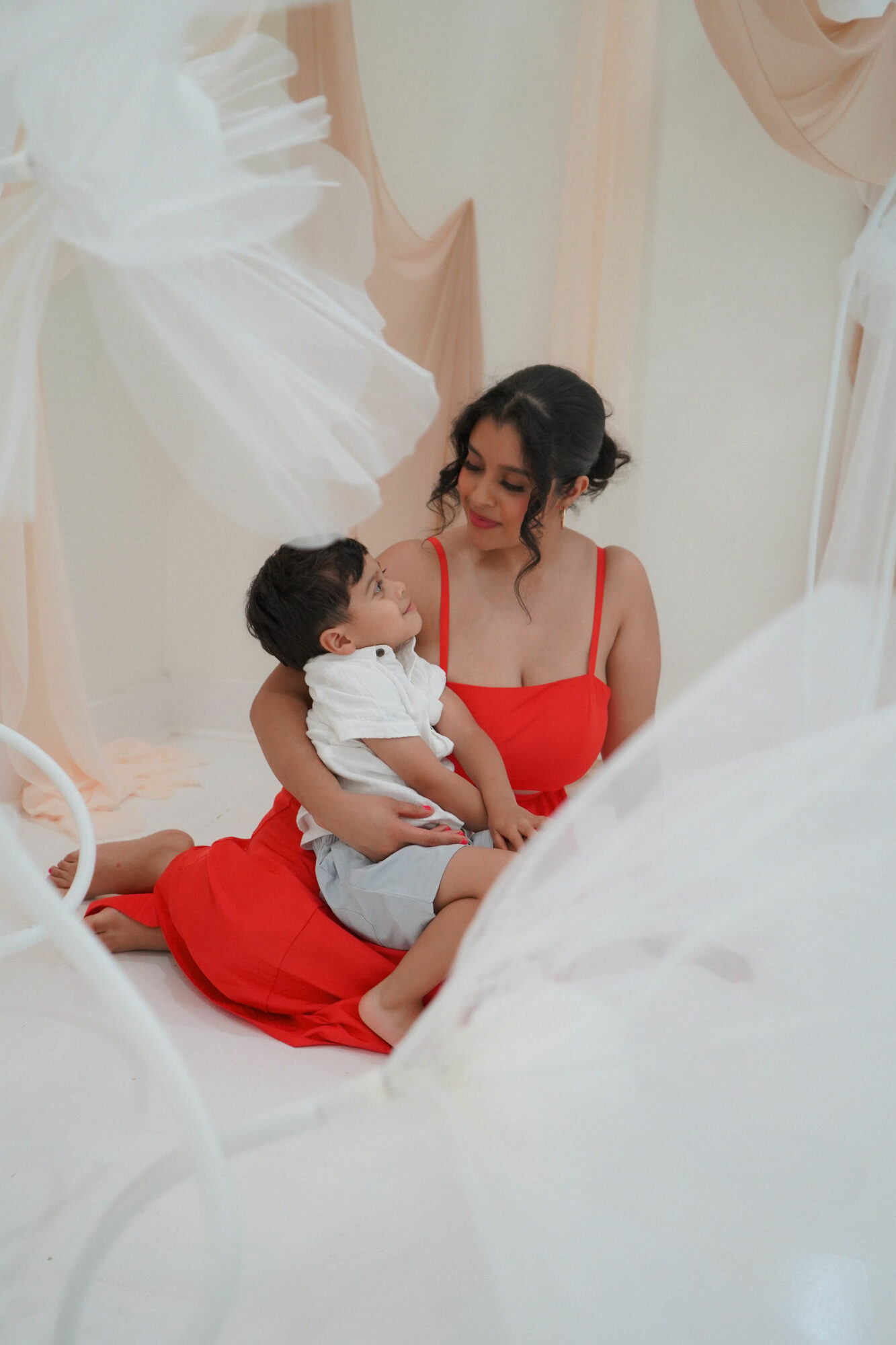 Woman in red dress sitting on floor holding a child, surrounded by white fabric and drapes.