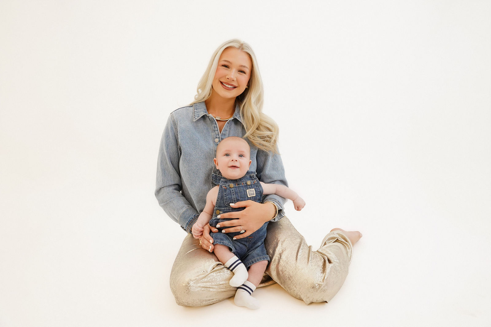 Woman sitting cross-legged holding a baby, both smiling, against a plain white background.
