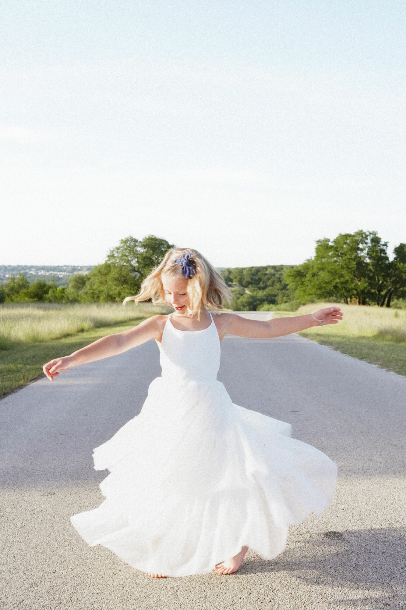 Woman in a white dress dancing on a road with trees and open sky in the background.