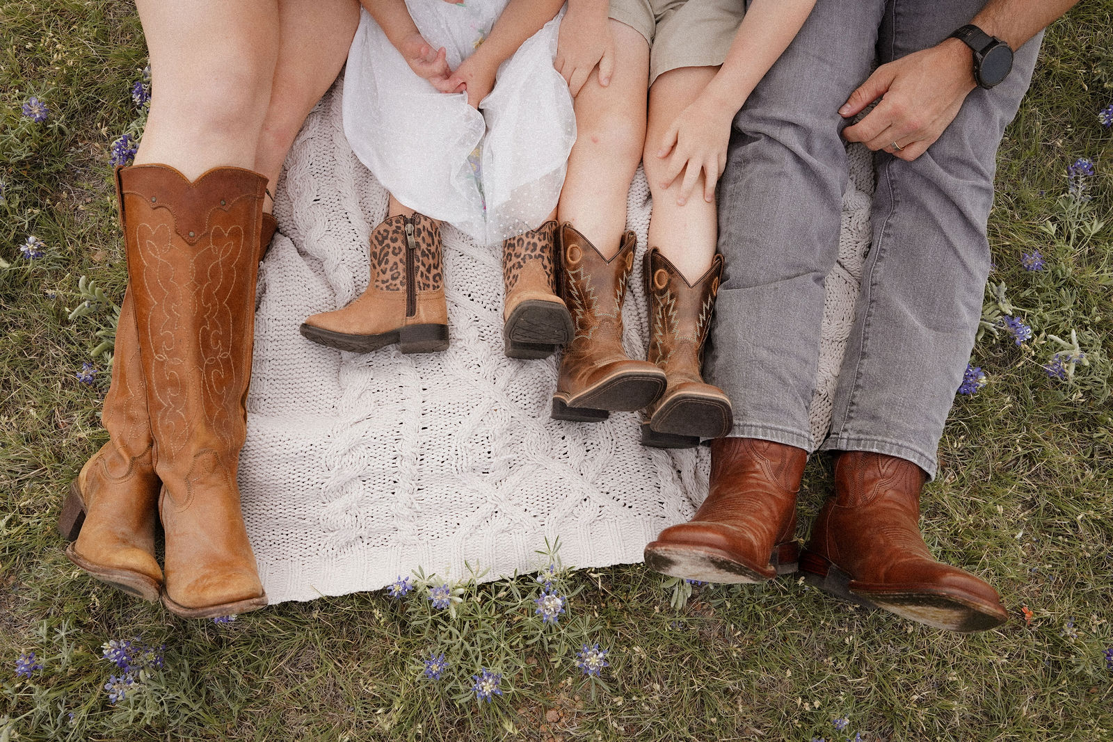 People sitting outdoors with their legs and boots visible, surrounded by grass and small purple flowers.