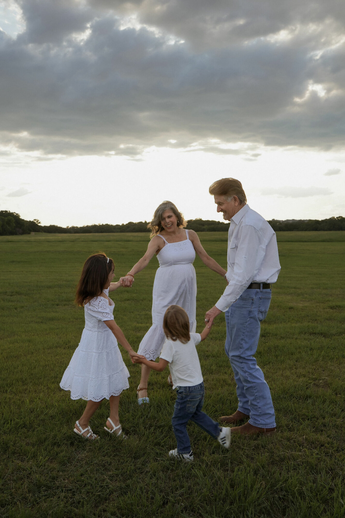 A family of four holding hands and dancing in a grassy field under a cloudy sky.