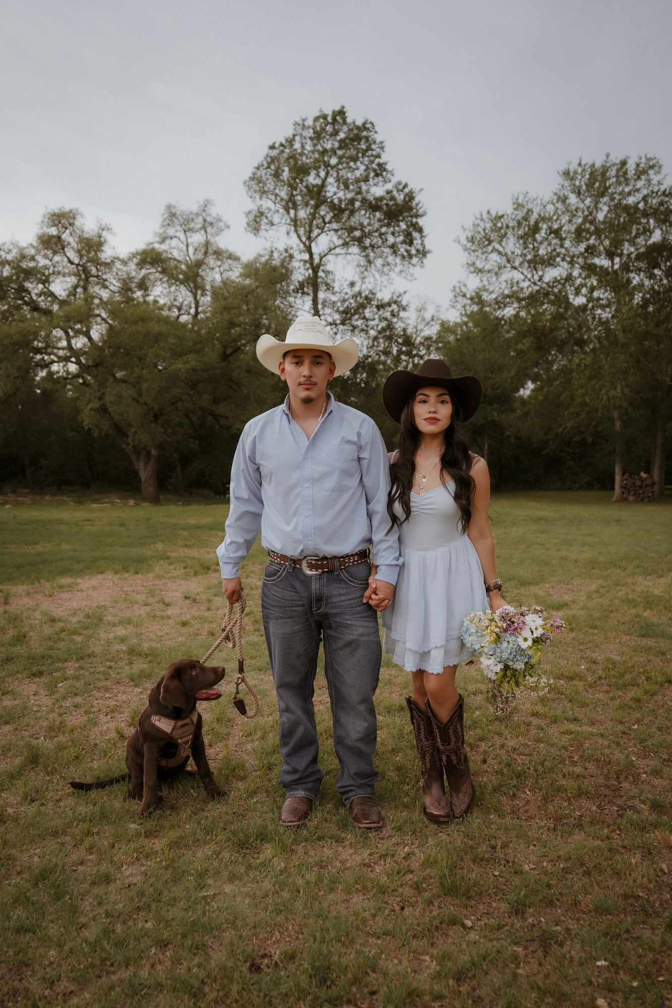 A couple holding hands with a dog in a park, trees in the background, overcast sky, woman holding a bouquet, both in cowboy hats.