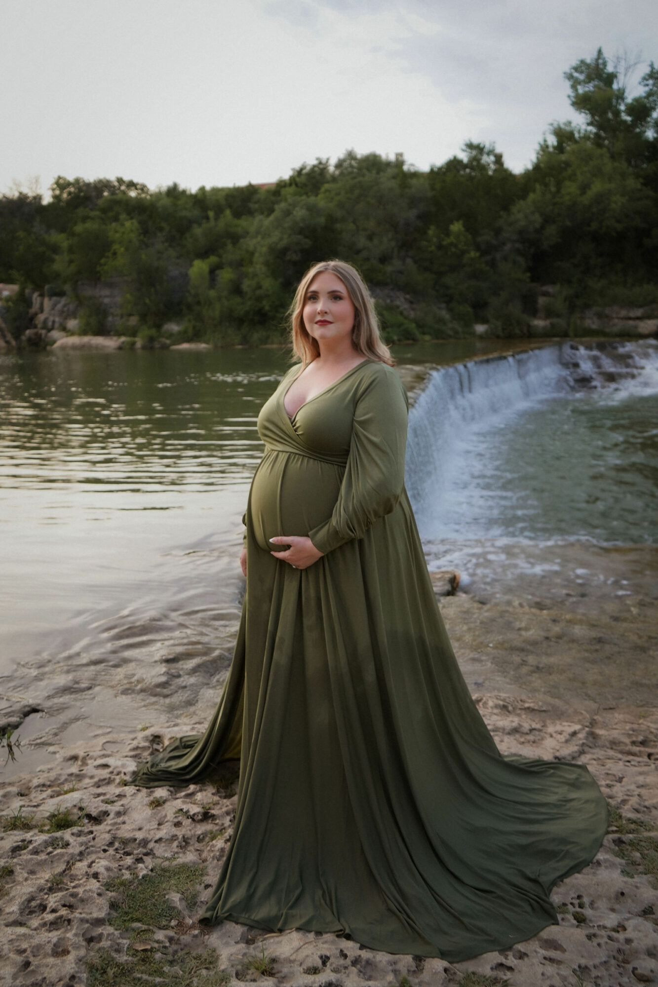 Pregnant woman in a long green dress standing by a river with a waterfall and trees in the background.