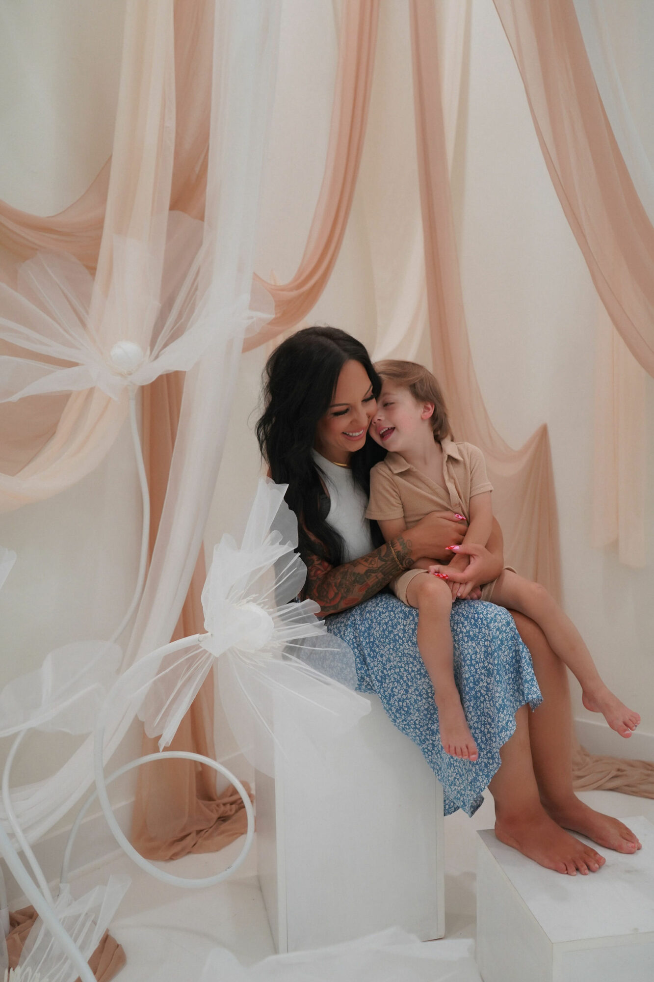 Woman and young girl sitting together, smiling, surrounded by soft, flowing curtains in a light-colored room.