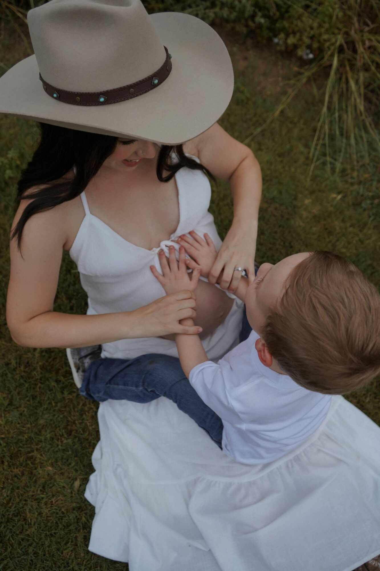 Woman wearing a large hat and white dress with a child outdoors on grass, holding hands.