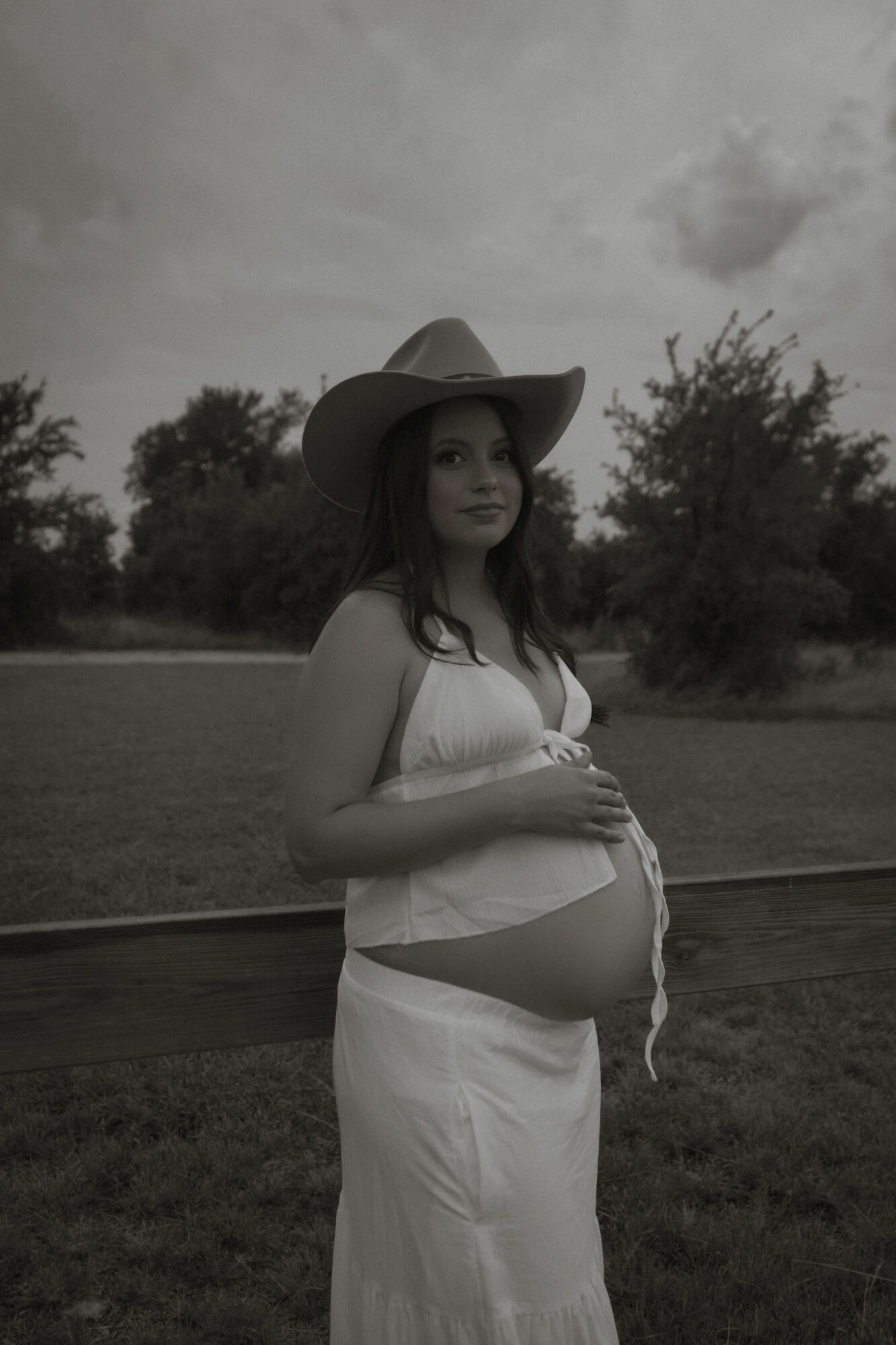 Pregnant woman wearing a wide-brimmed hat and white dress outdoors, holding her belly, with trees and cloudy sky in background.