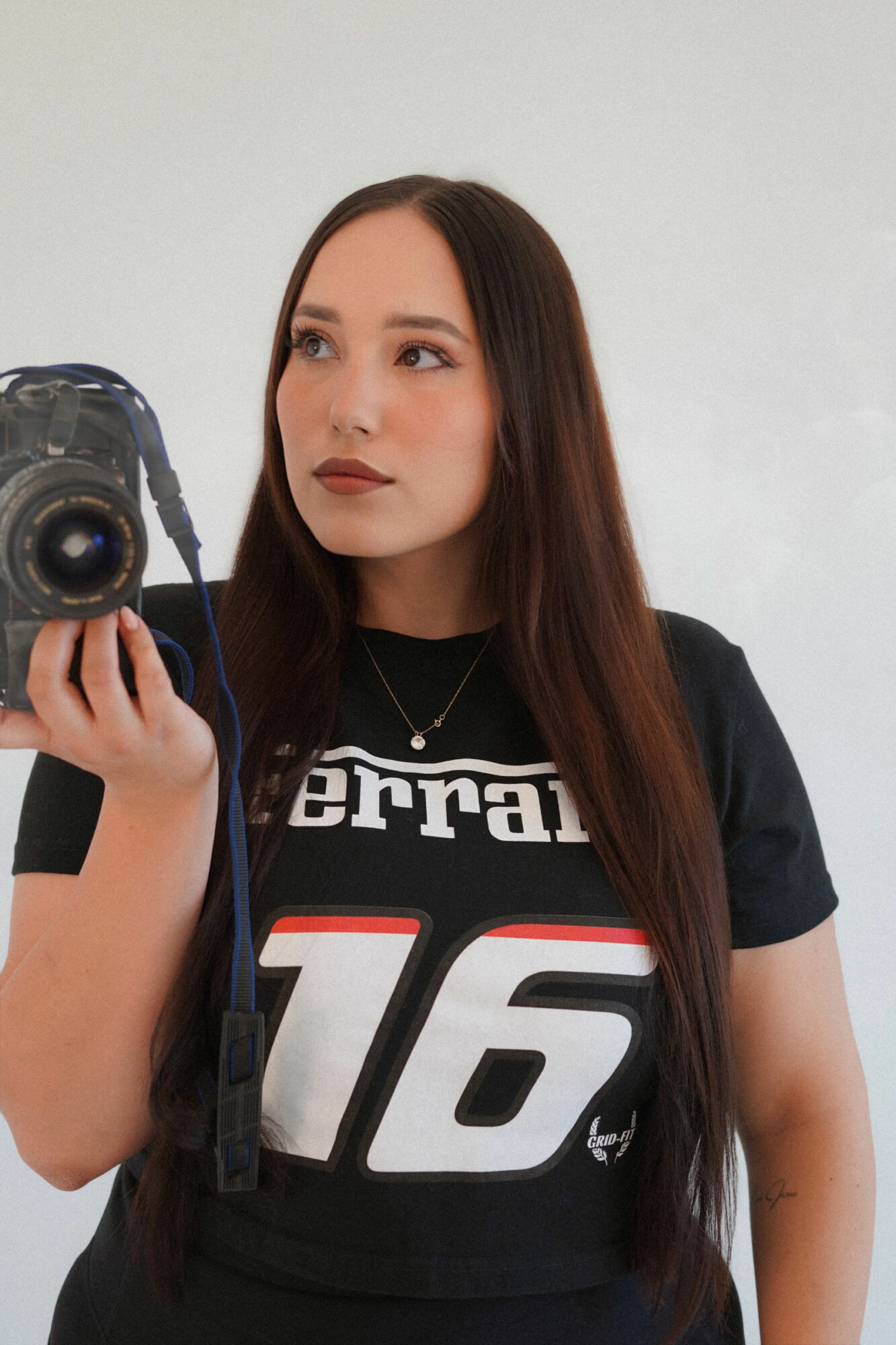 Young woman with long brown hair taking a selfie with a camera, wearing a black sports jersey with the number 16.
