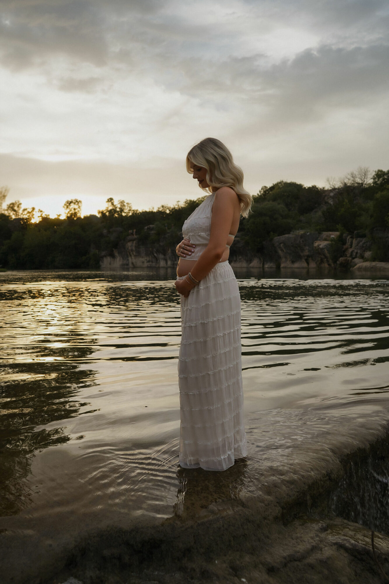 Woman standing in water near shoreline, looking down, with trees and cloudy sky in background.