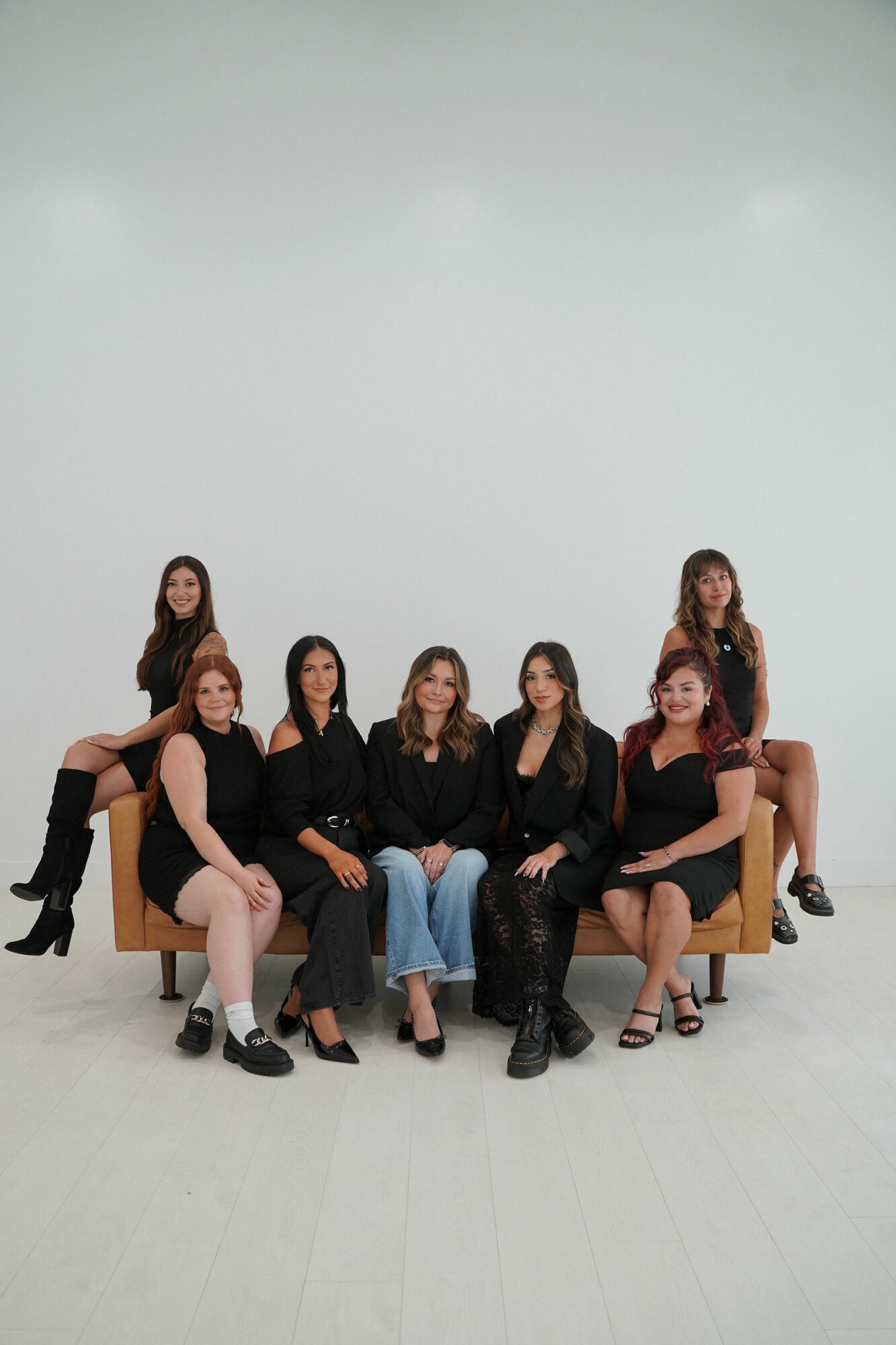 Six women sitting and standing around a sofa in a studio with a plain background.
