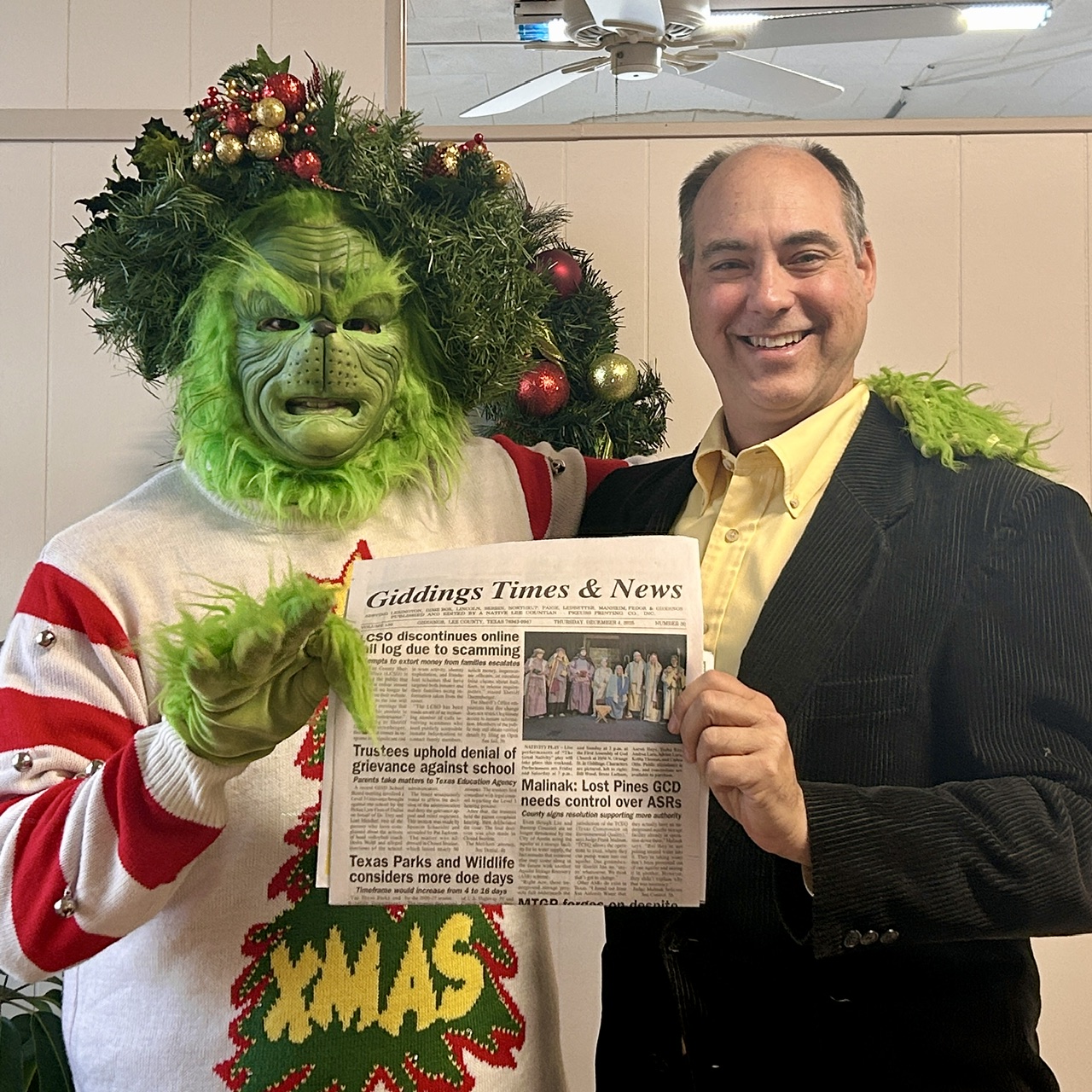 Person in a green lion costume and a man in a suit hold a newspaper together, smiling. The costume has a large green face and mane, with a red and white striped shirt. The man wears a dark suit and yellow shirt. They are indoors with a ceiling fan visible. The newspaper headline reads
