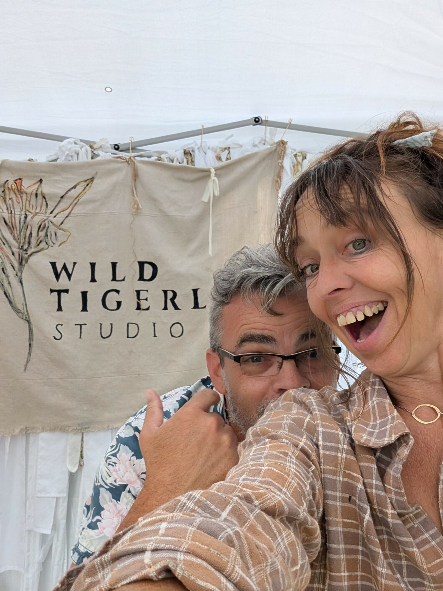 Two people smiling and taking a selfie in front of a banner that reads 'WILD TIGERL STUDIO'.