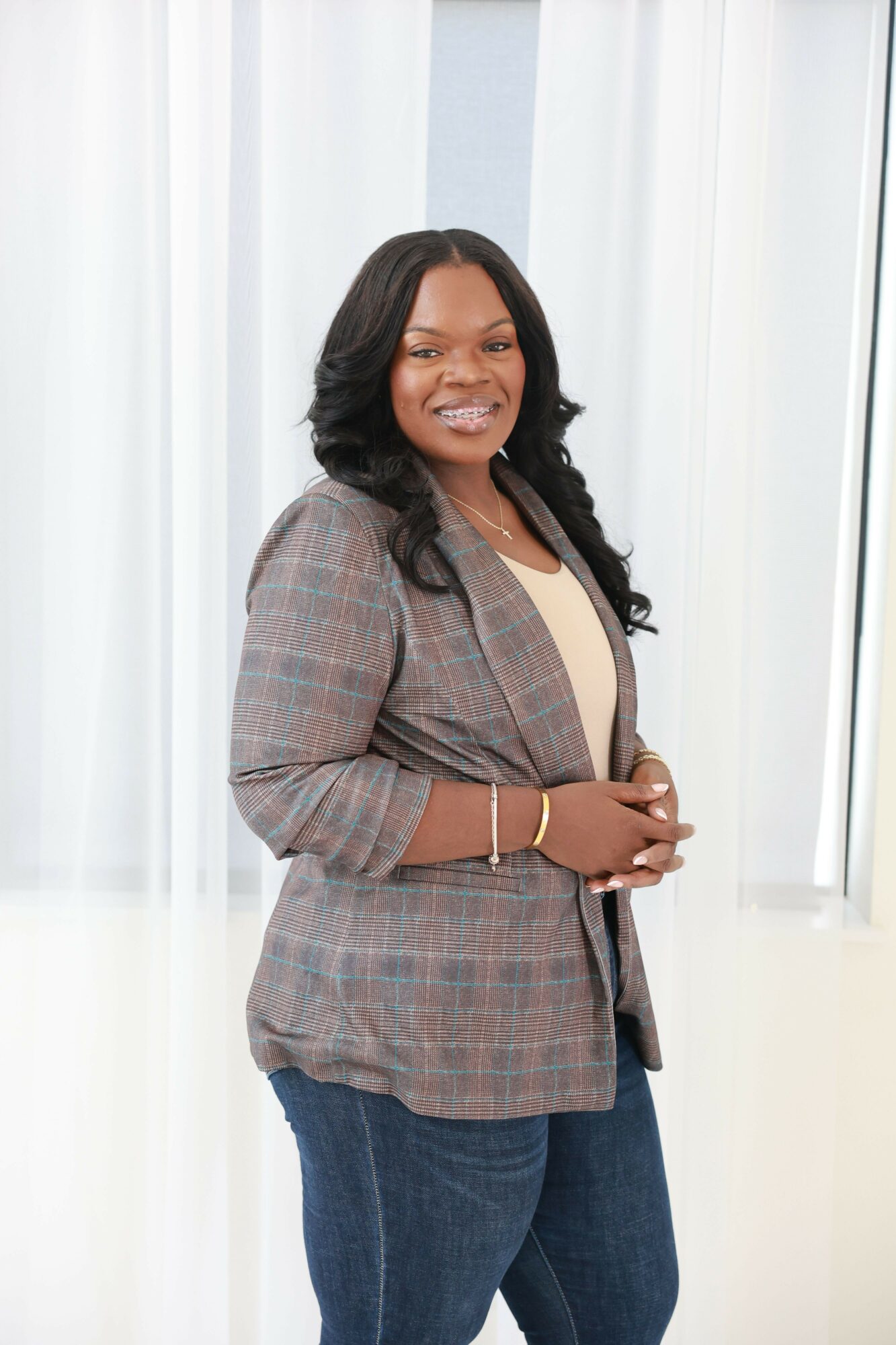 Woman standing indoors, smiling, with hands clasped in front, wearing a plaid blazer and dark pants.